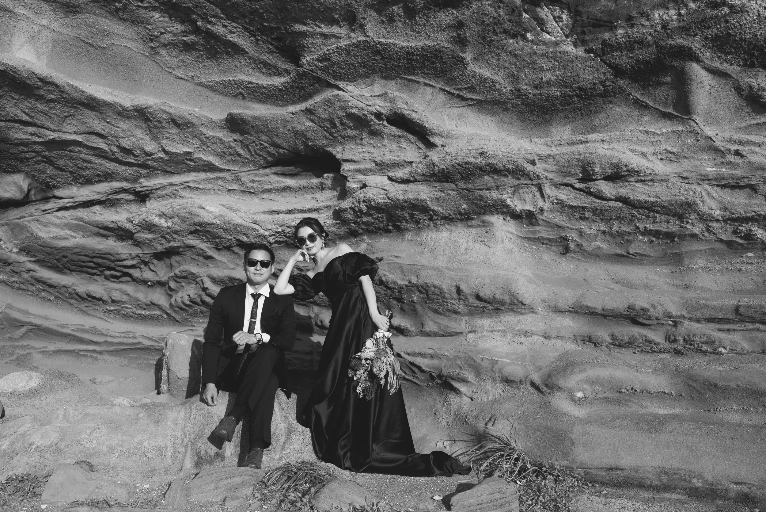 サングラスをかけた男性が座り、女性は長いドレスを着て花束を持ち、岩場に立っている。