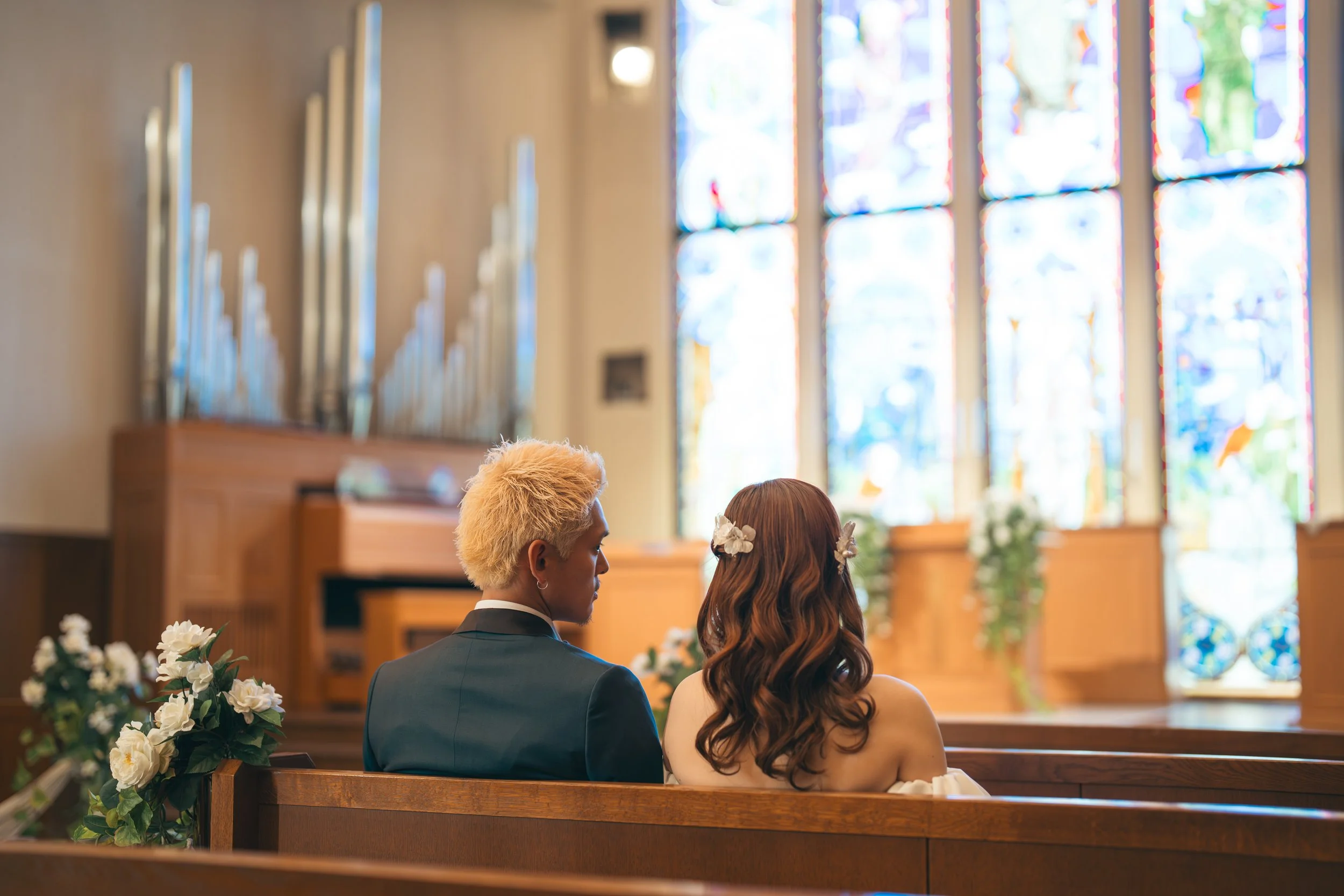 教会で結婚式を挙げているカップル。男性は黒いスーツ、女性は白いドレス。背景にステンドグラスとパイプオルガンが見えるです。