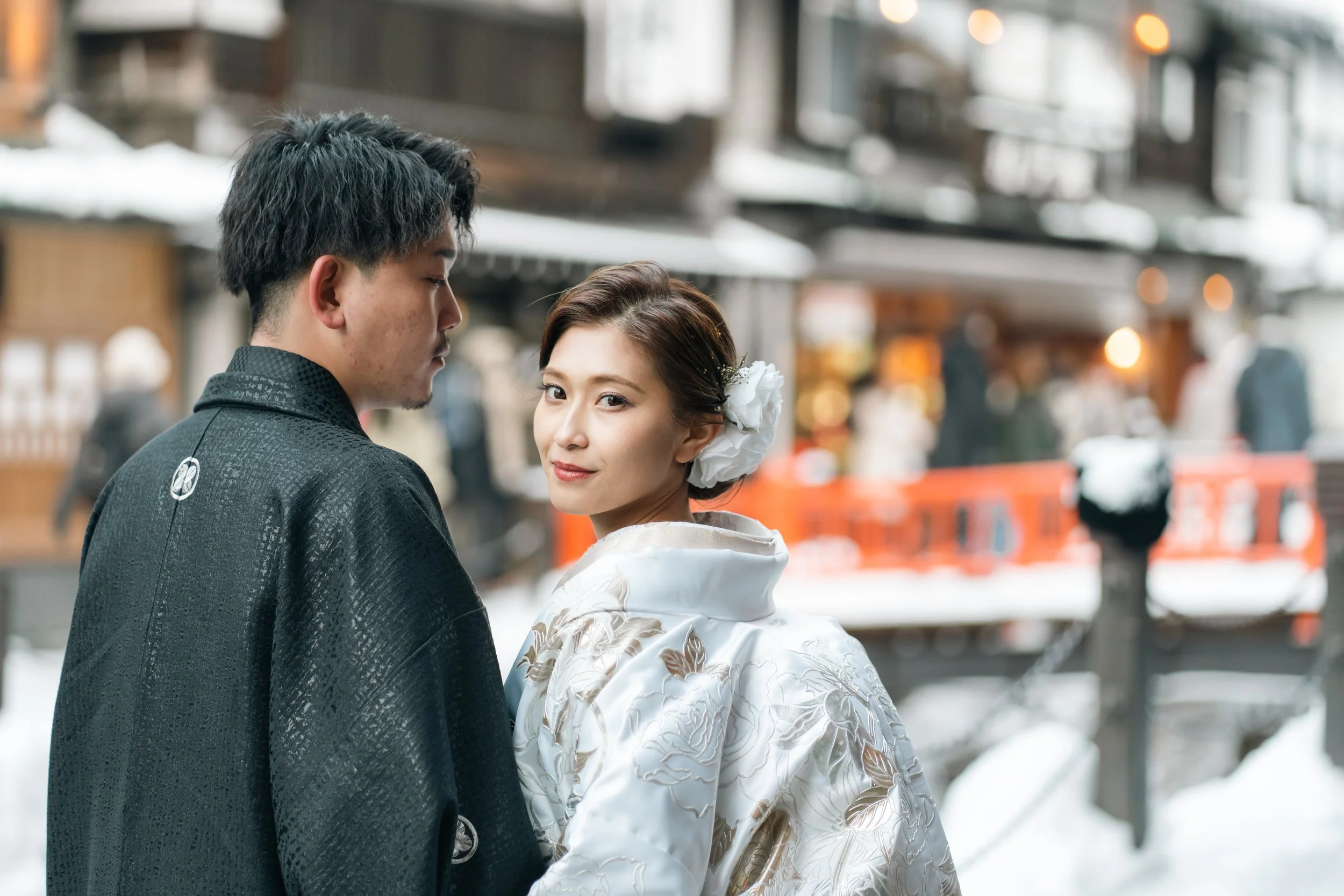 雪の積もった町並みの中で、伝統的な和装を着た夫婦が立っている。女性は白の着物に花飾りをつけている。