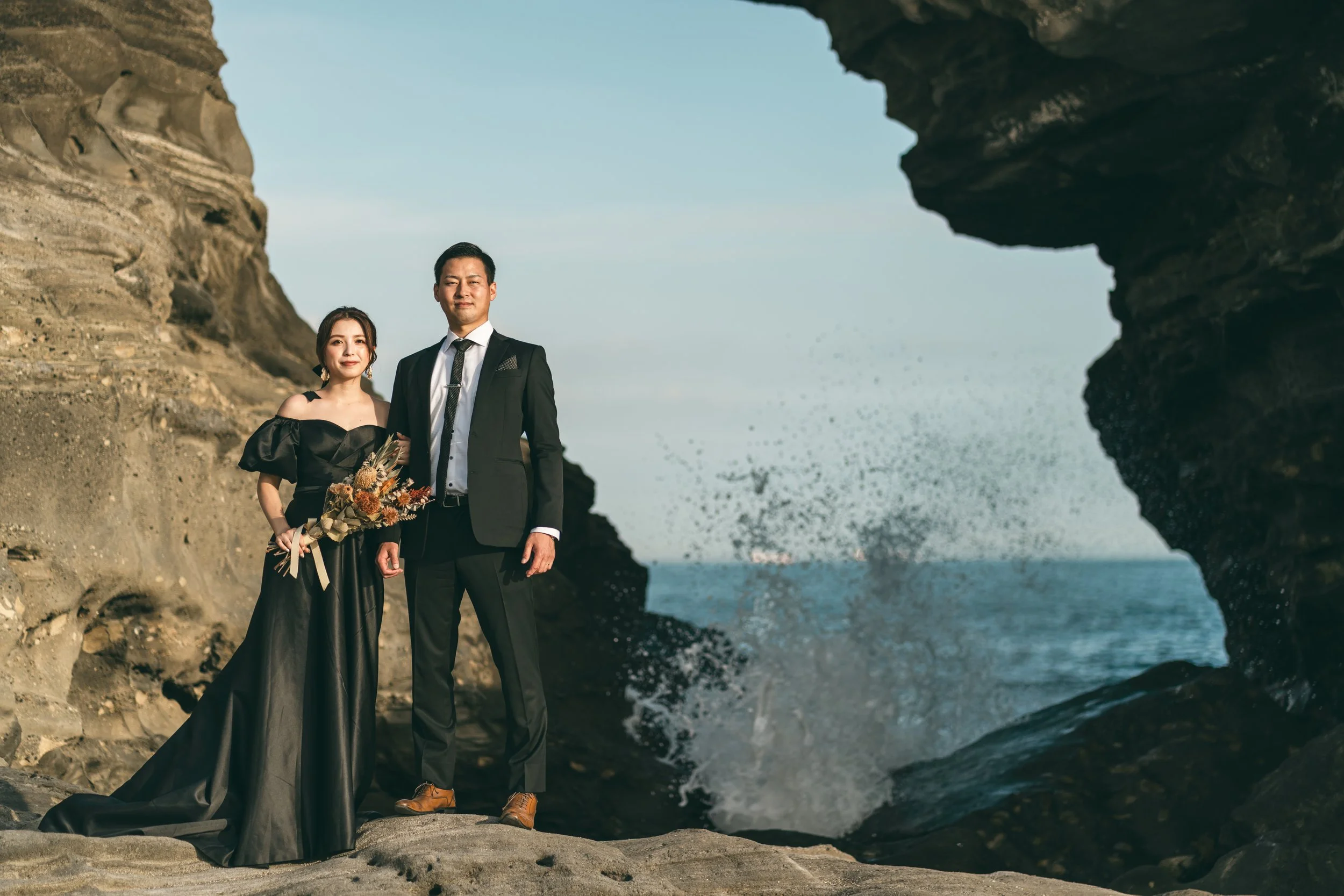 海の近くの岩場に立つ夫婦のカップル。女性は黒いドレスを着て花束を持ち、男性は黒いスーツを着ている。背景には海と青空が広がる。