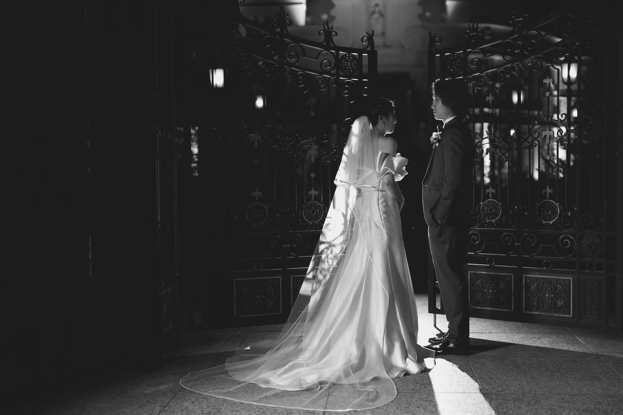 夜の光の中で結婚式の新郎新婦がゲートの前で見つめ合っているシーン。二人はフォーマルな服装で、女性は長いドレスとヴェールを着用している。