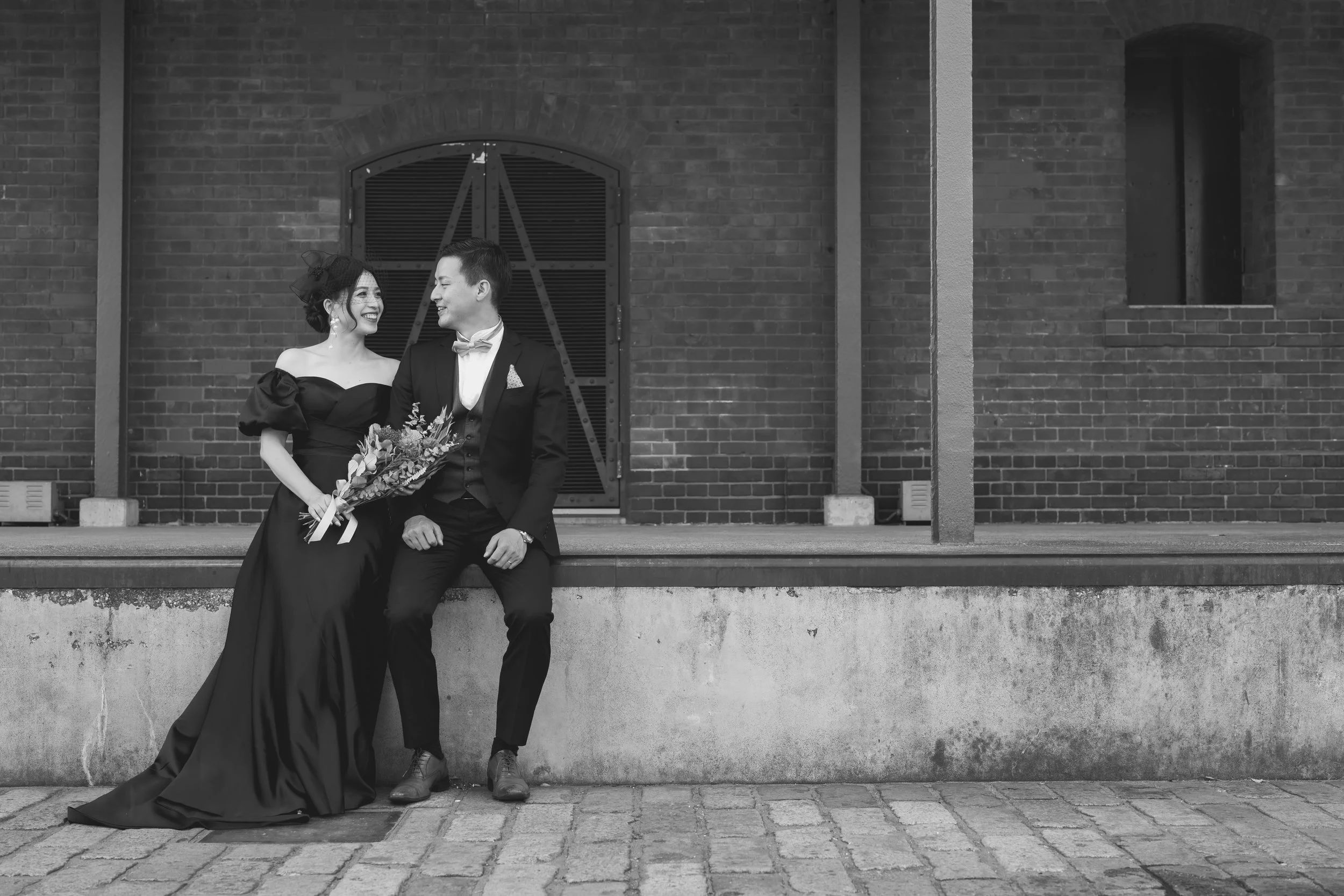 結婚式の写真。黒いドレスを着た女性とスーツを着た男性がベンチに座り、お互い笑顔で見つめ合っている。女性は花束を持っている。背景はレンガ造りの建物。