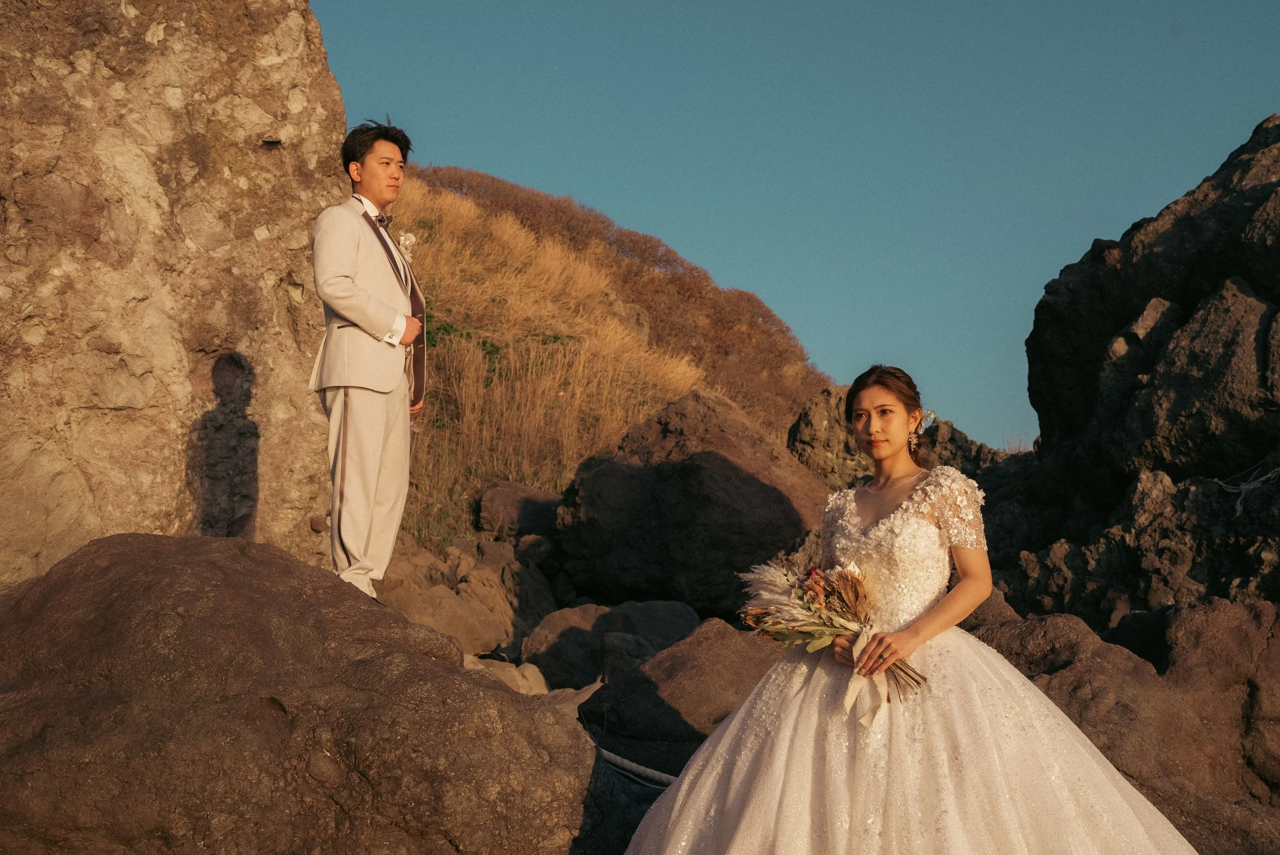 山の岩場でウェディング写真を撮影しているカップル。女性は白いドレス、男性はグレーのスーツを着ている。夕日の光が二人を照らしている。