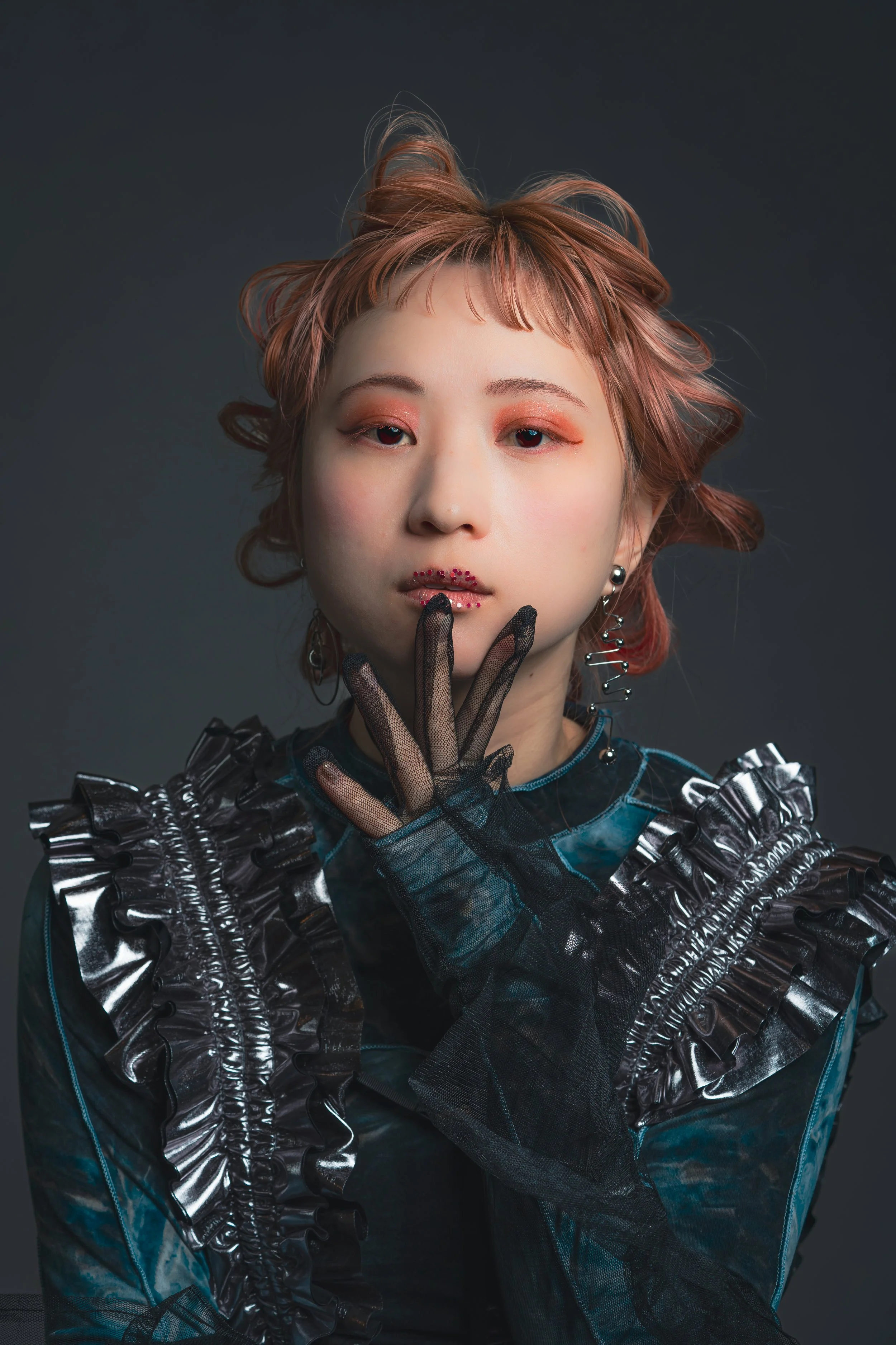 暗い背景に、ピンク色の髪の女性が黒いレースの手袋をつけ、ファッショナブルな衣装を着てポーズをとっている写真。