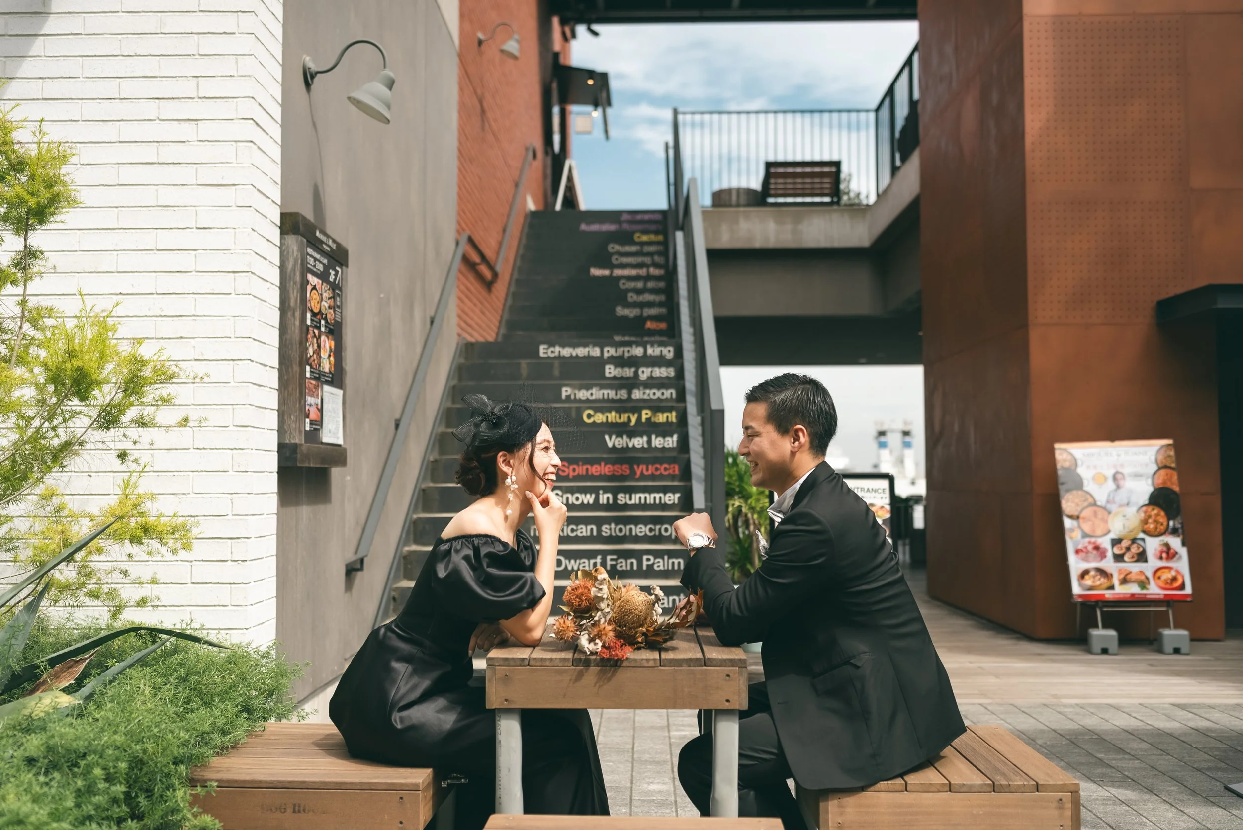 屋外のレストランで男女が笑顔で会話しているシーン。テーブルには花束が置かれている。背景に階段とメニュー表がある。
