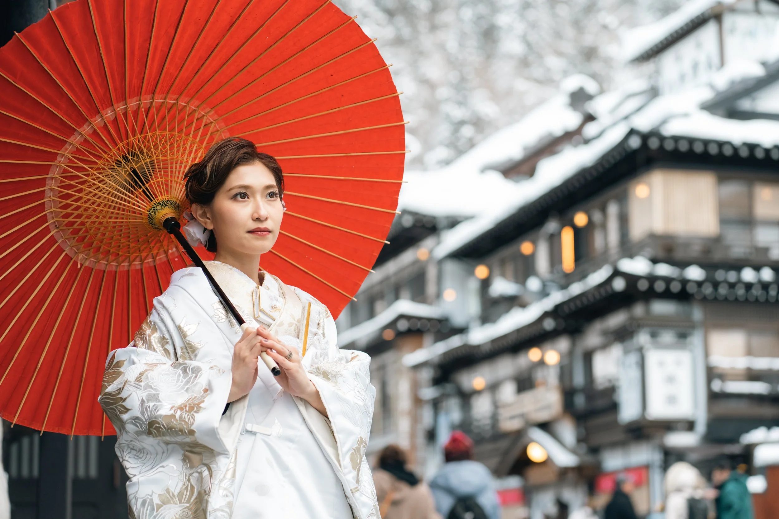 雪の中の伝統的な日本の町並みの前で、白い着物を着た女性が赤い和傘を持っている。