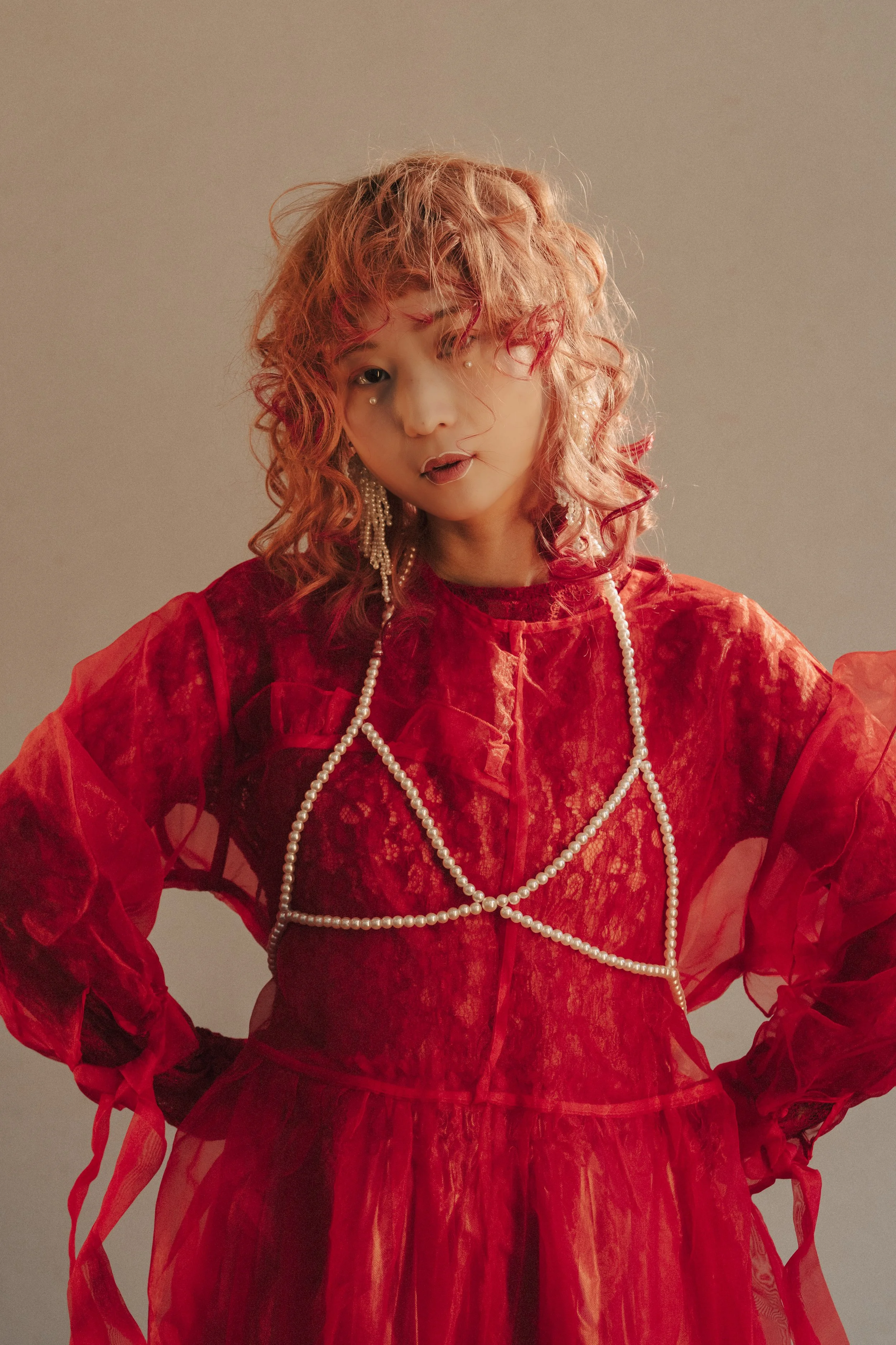 赤いレースのドレスを着た女性が中央に立ち、肩に手を置いている。髪はカールし、赤みがかった色合い。イヤリングと長いパールのネックレスをつけている。