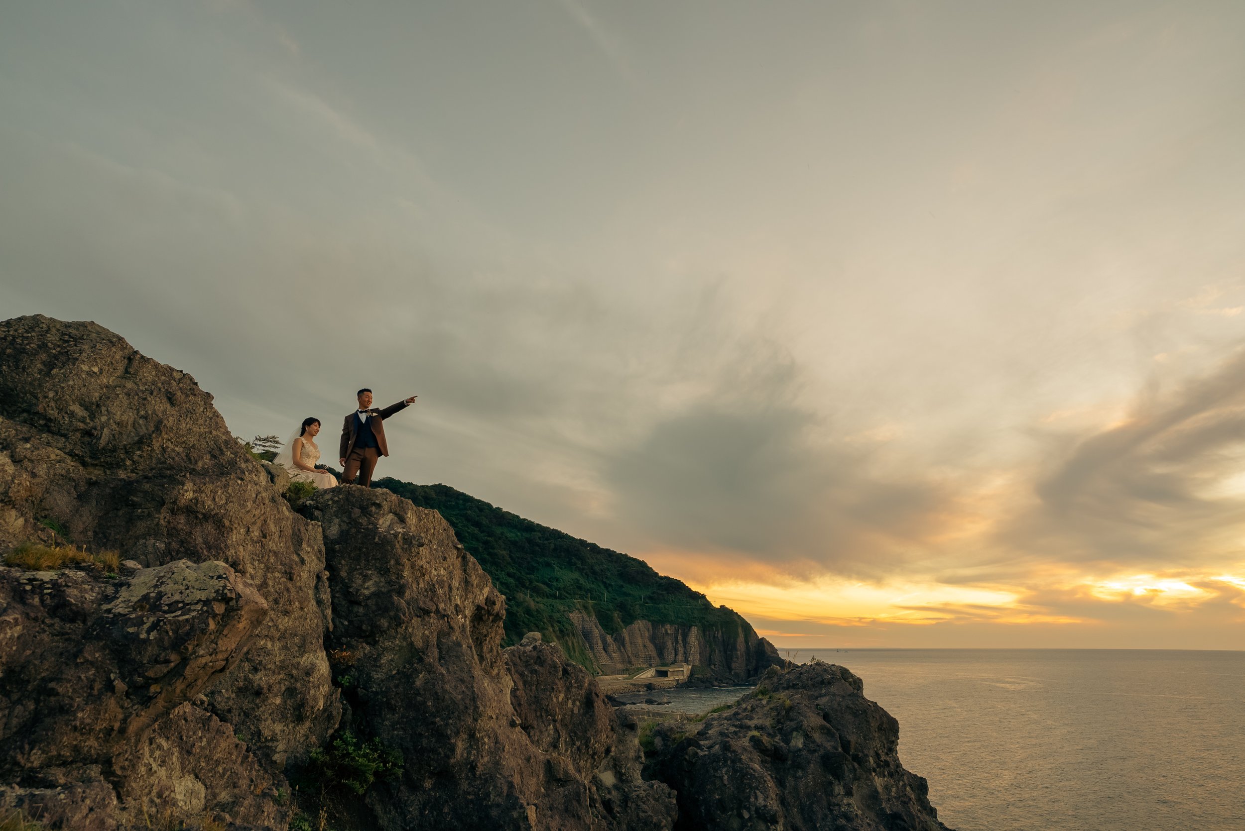 夕焼けの海辺の崖の上に立つ男女。男性はスーツを着て指をさし、女性はドレス姿で座っている。