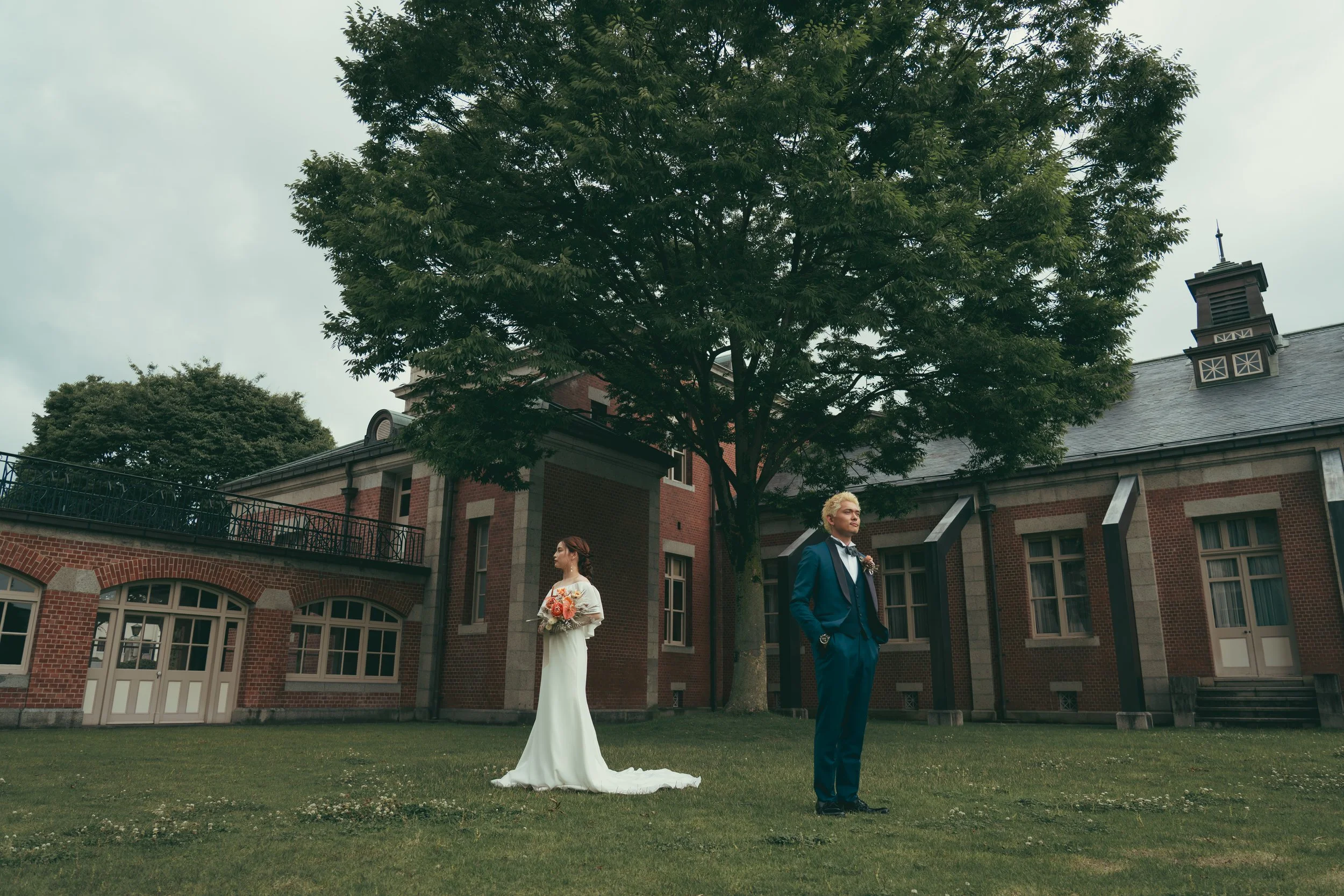 結婚式のカップルが屋外の庭で立っている。女性は白いドレスを着て花束を持ち、男性は青いスーツを着ている。大きな木の下に背を向けて歩いている。背景には赤レンガの建物と曇り空が見える。