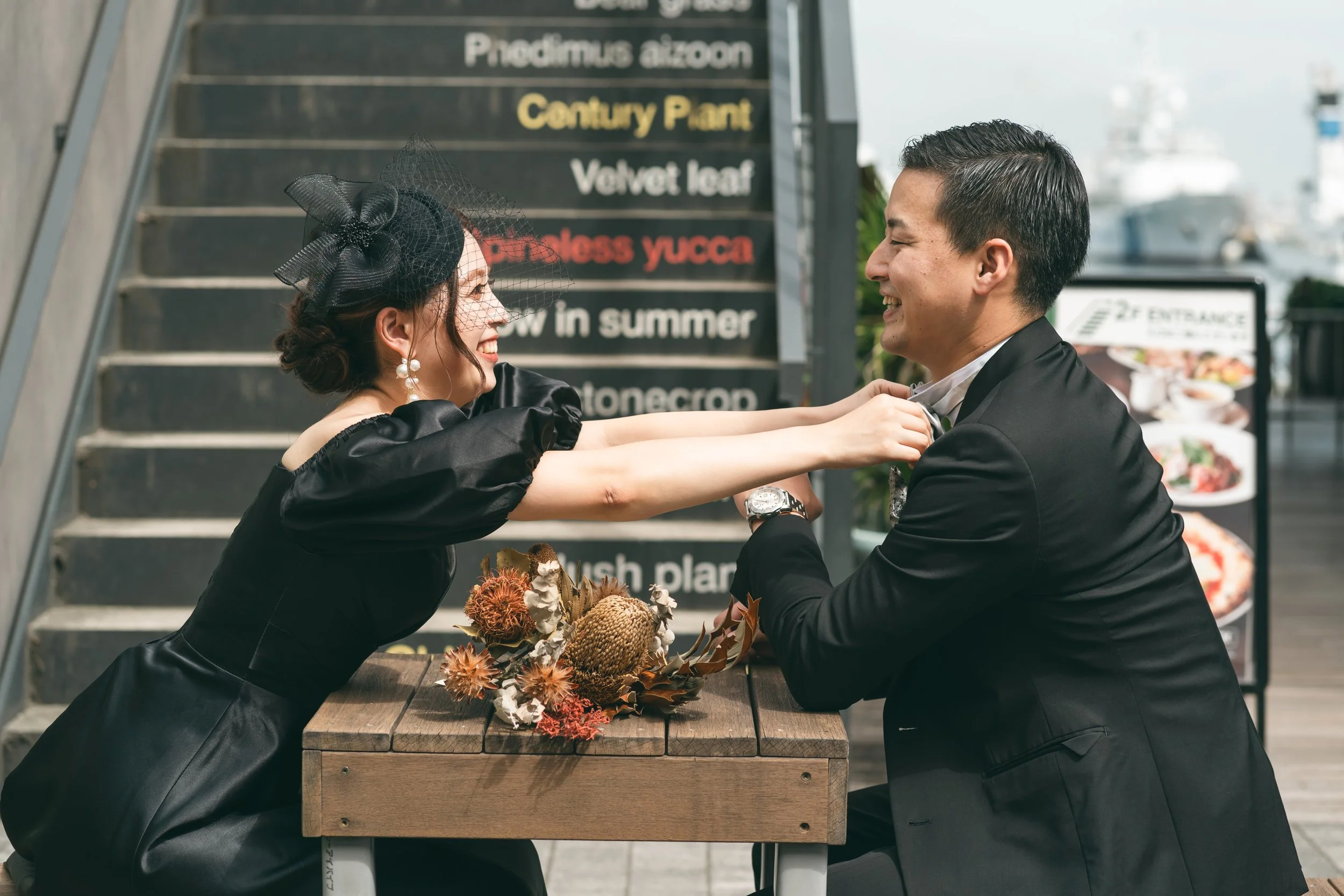 男性と女性がレストランの外のテーブルで結婚式の誓いを交わしている。女性は黒いドレスと帽子を身に着けている。男性は黒いスーツを着ている。女性が男性のネクタイを結んでいる。テーブルには花束がある。