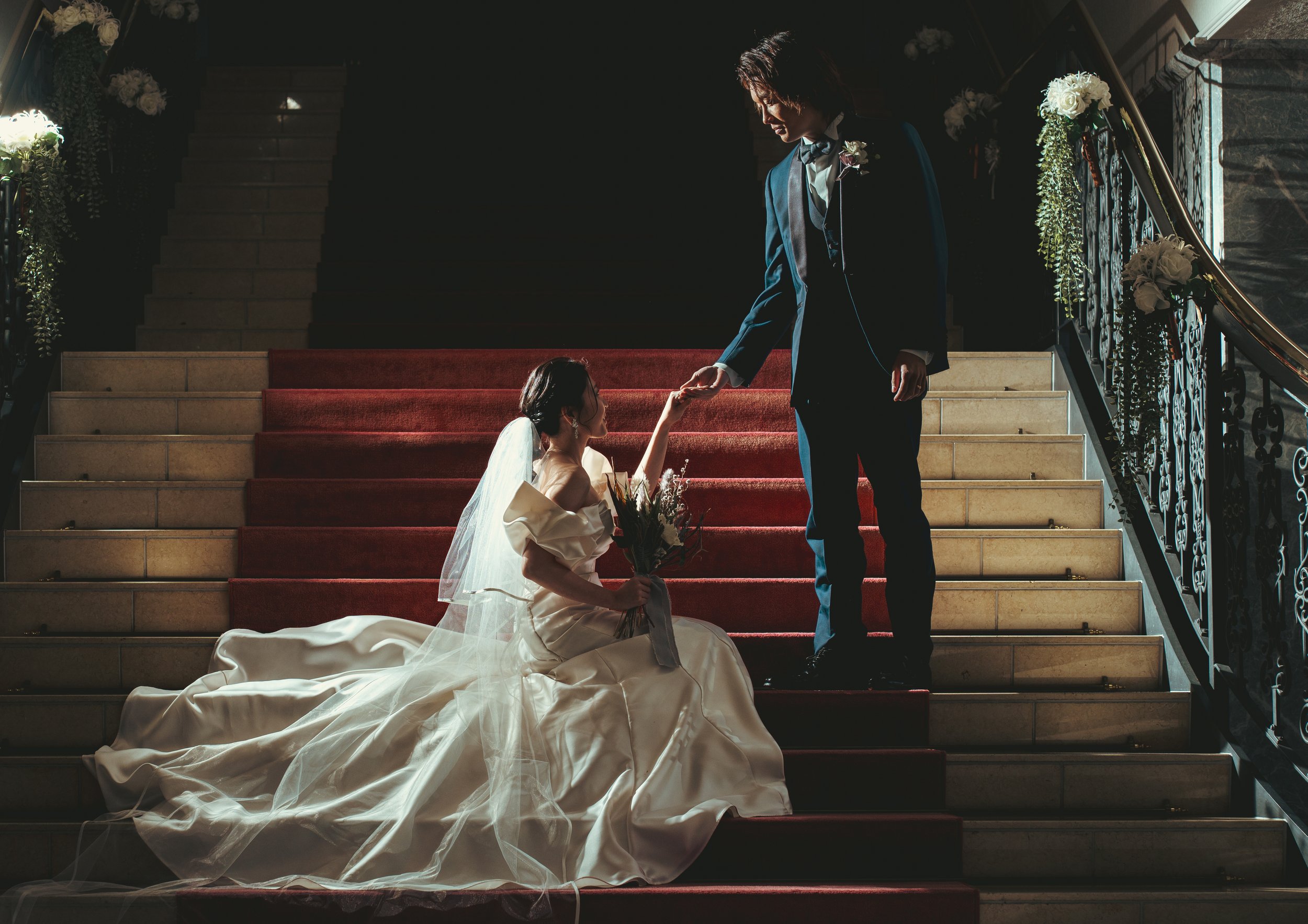階段の上で結婚式を迎えるカップル、女性は白いドレスとヴェールを着ていて、男性は黒いスーツと蝶ネクタイを着ている。女性は花束を持ち、男性は彼女の手を取っている。
