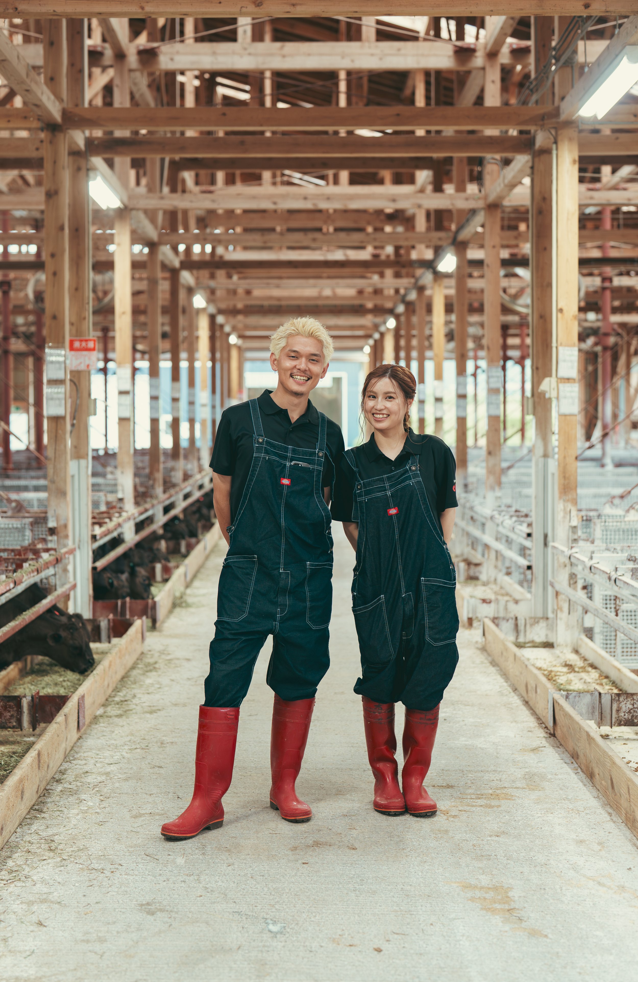 牛舎で働く二人の若い男女スタッフ、黒い作業服と赤い長靴を着用し、笑顔で並んでいる