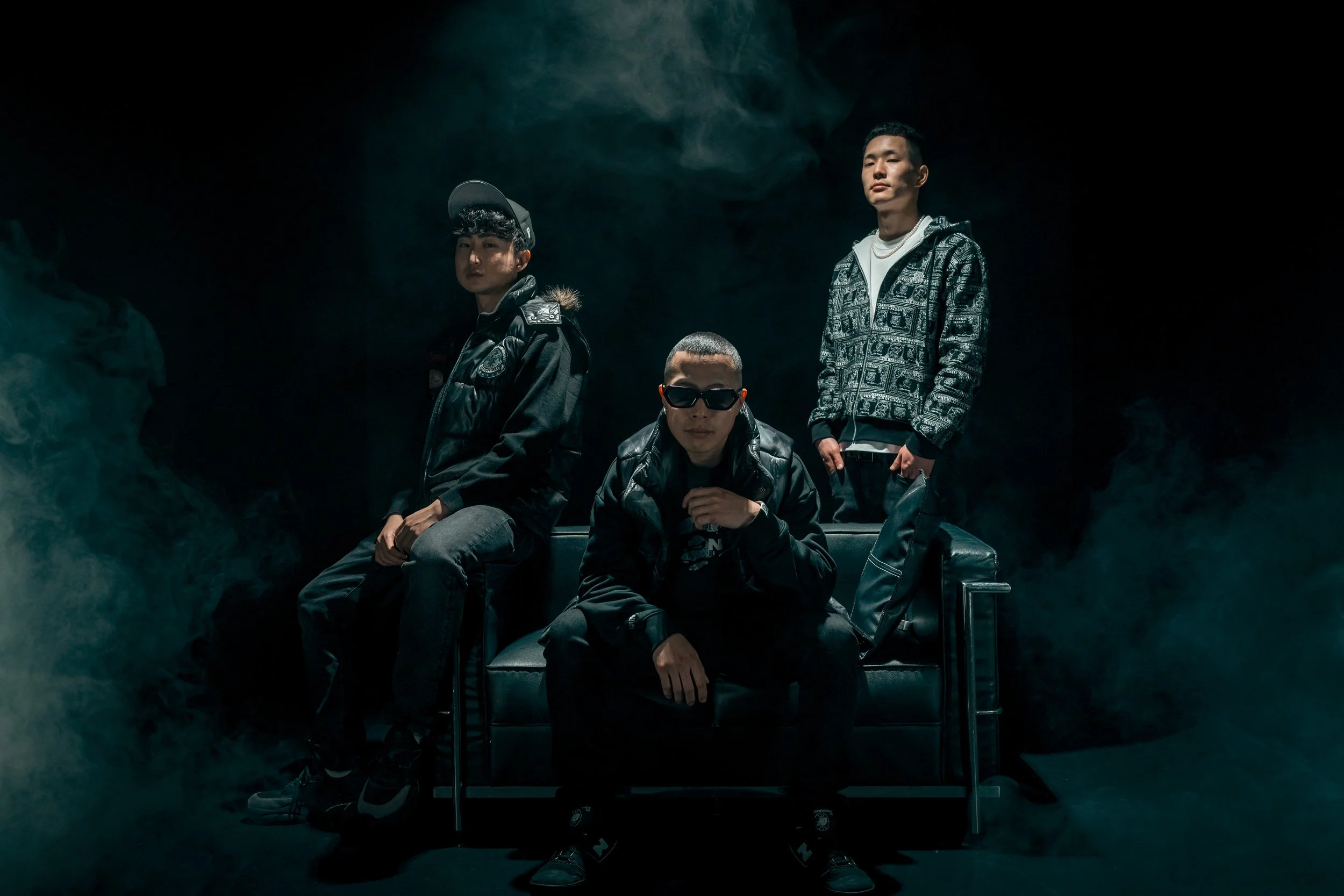 黒背景に立つ三人の男性のグループ写真。中央の男性はサングラスをかけ、革ジャンを着ている。左と右の男性はカジュアルな服装で立っている。暗い照明と煙の演出が雰囲気を作り出している。