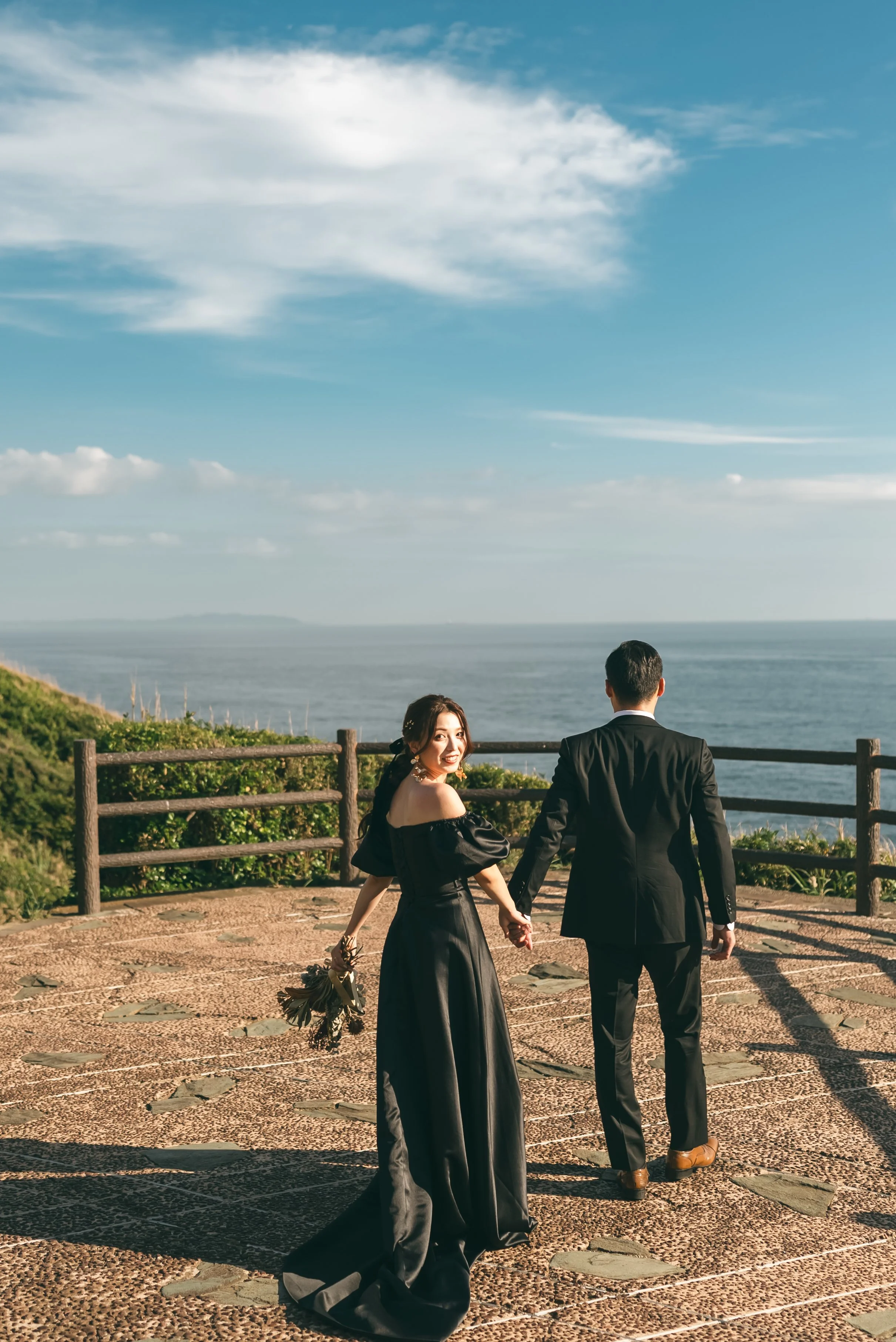 海沿いの展望デッキで手をつないで歩くカップル。女性は黒のドレスと花束、男性はブラックスーツ。青い空と海、緑の丘が背景。
