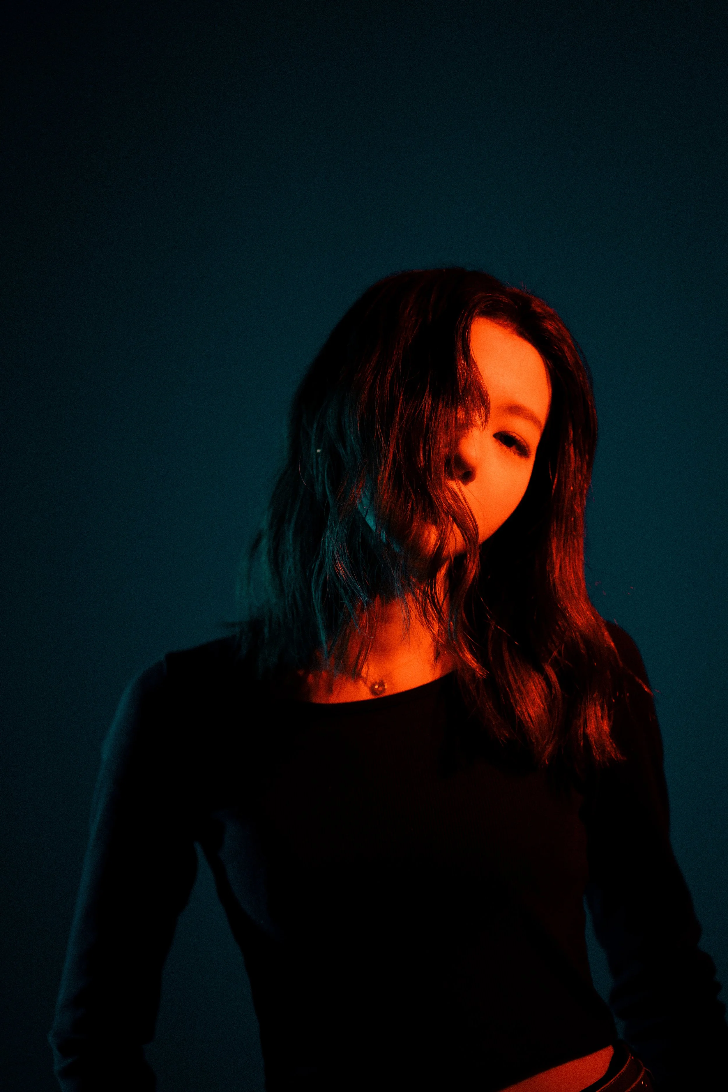 暗い背景に照らされた女性のポートレート。彼女の髪は肩まで伸びていて、顔の一部が隠れている。