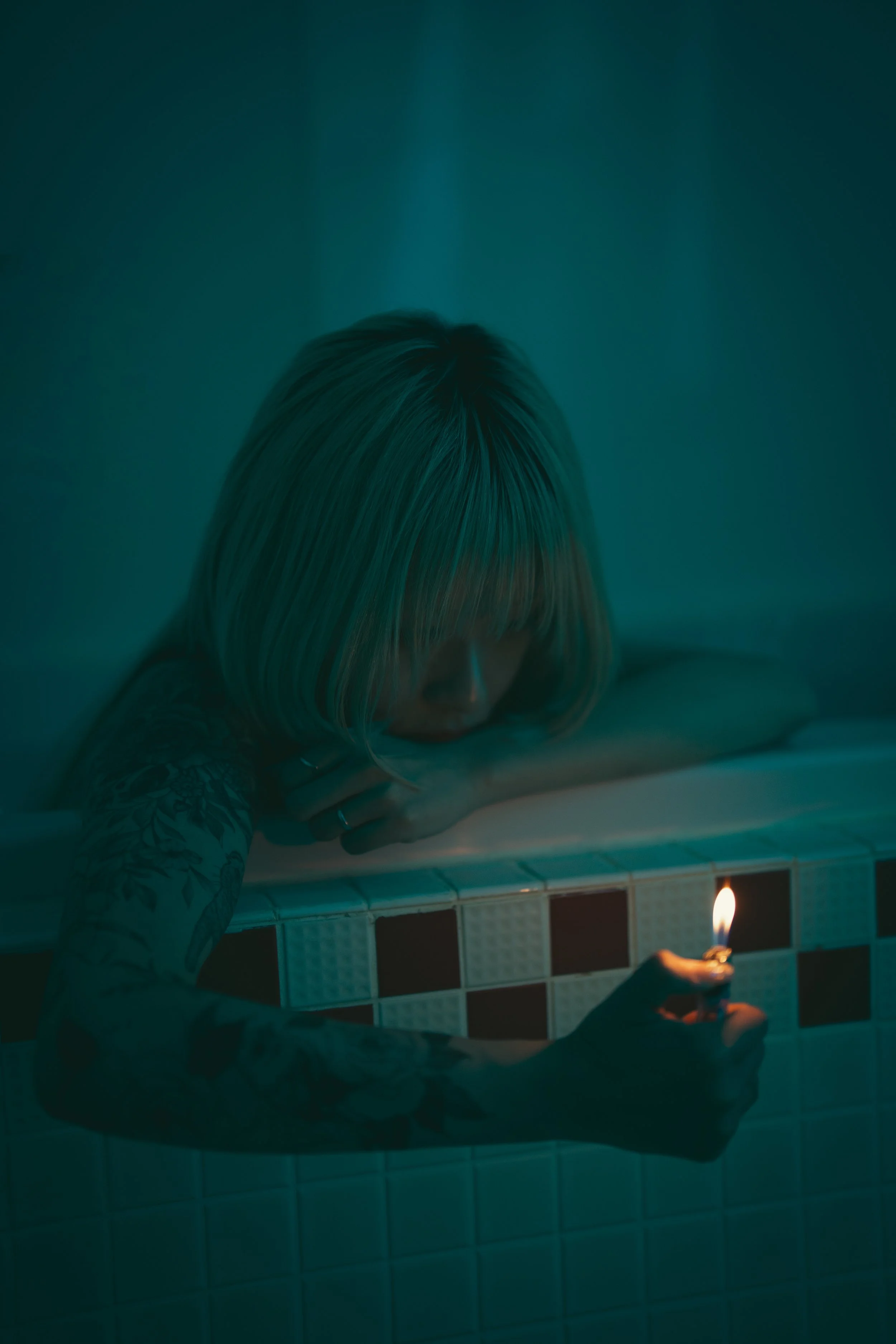 バスタブの縁に頭をのせて、キャンドルの火を持ち、暗い部屋の中にいる女性。