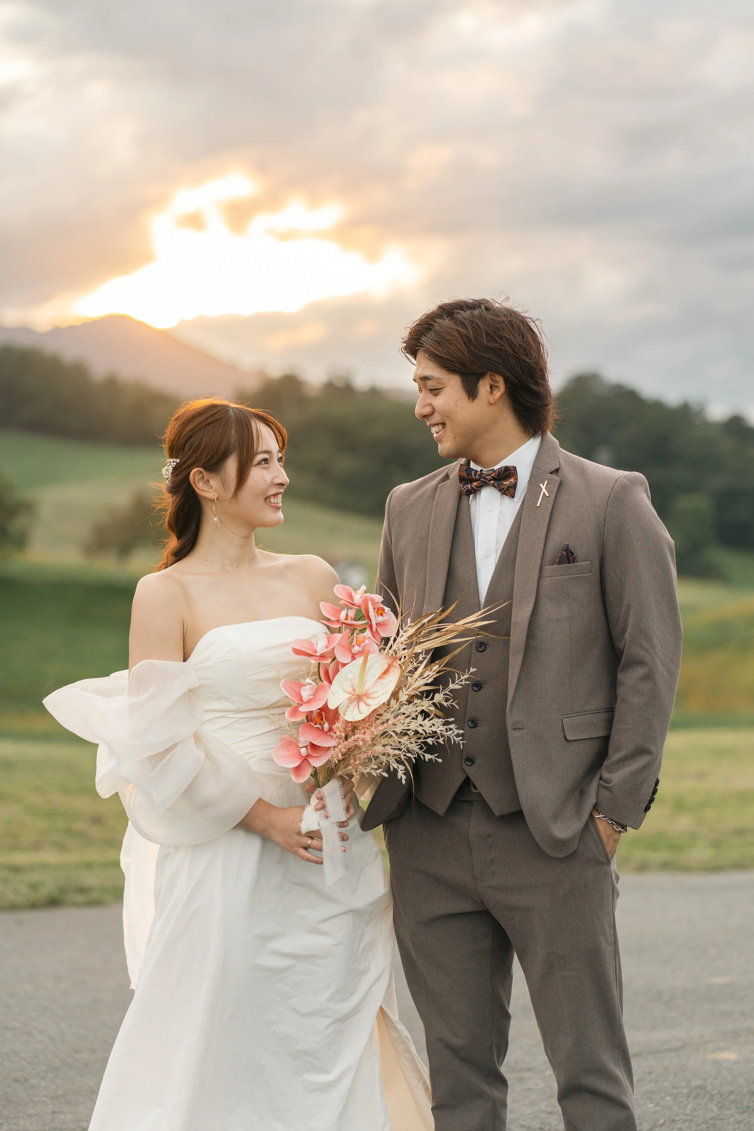 結婚式のカップルが笑顔でお互いを見つめ合っている風景。背景に夕焼けの空と自然の風景が広がっている