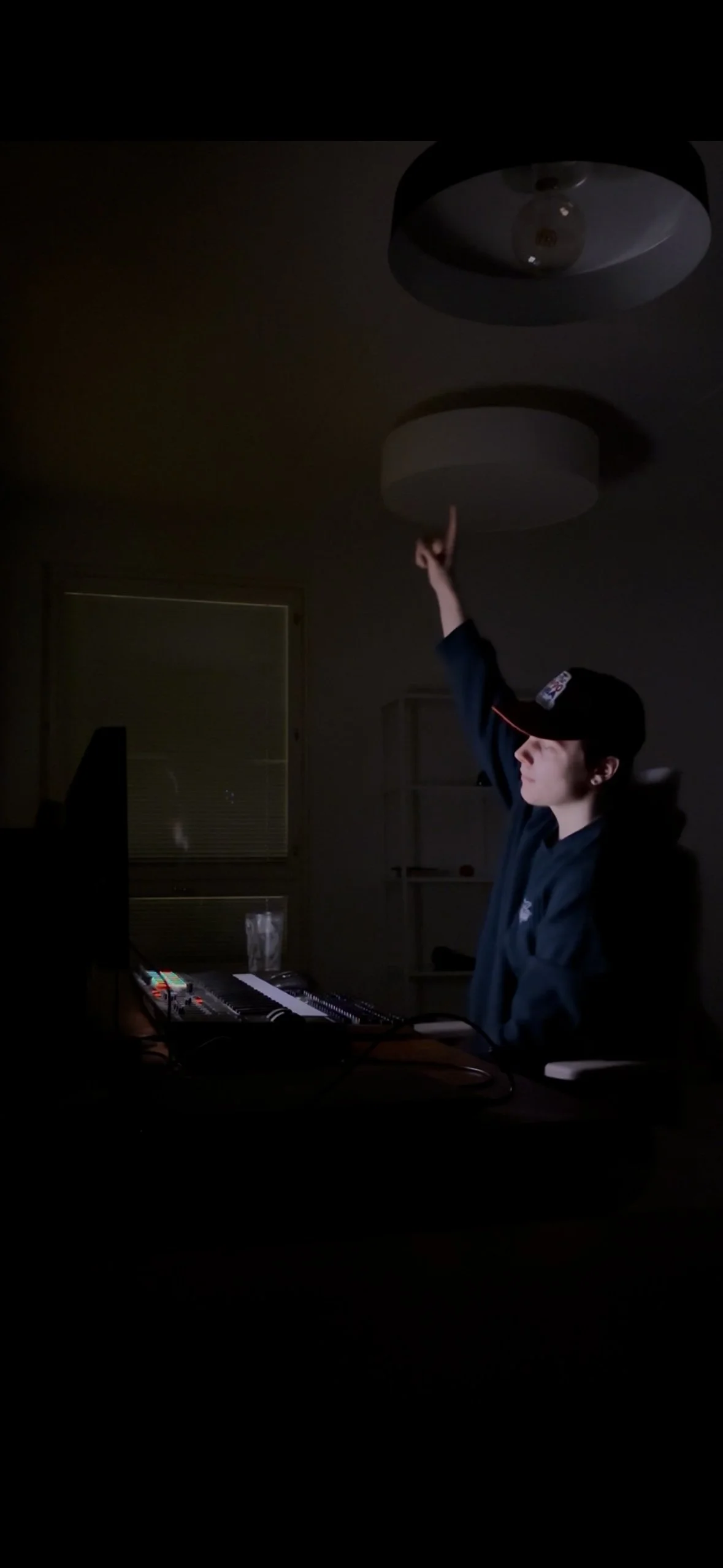 A young man sitting at a desk in a dark room, illuminated by computer and keyboard lights, reaching up towards a ceiling light with his right hand.