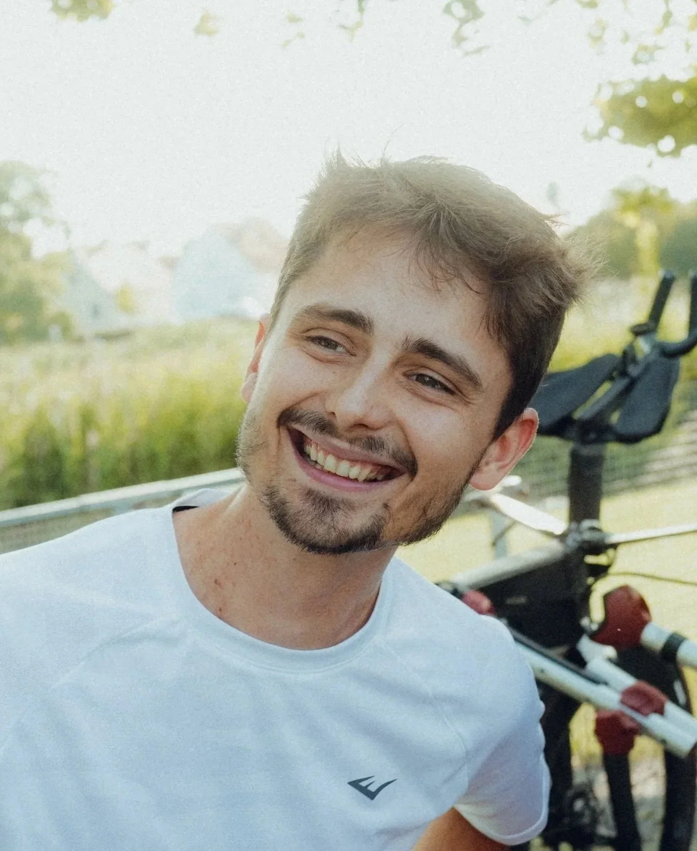 A young man smiling outdoors in front of a bicycle on a sunny day.