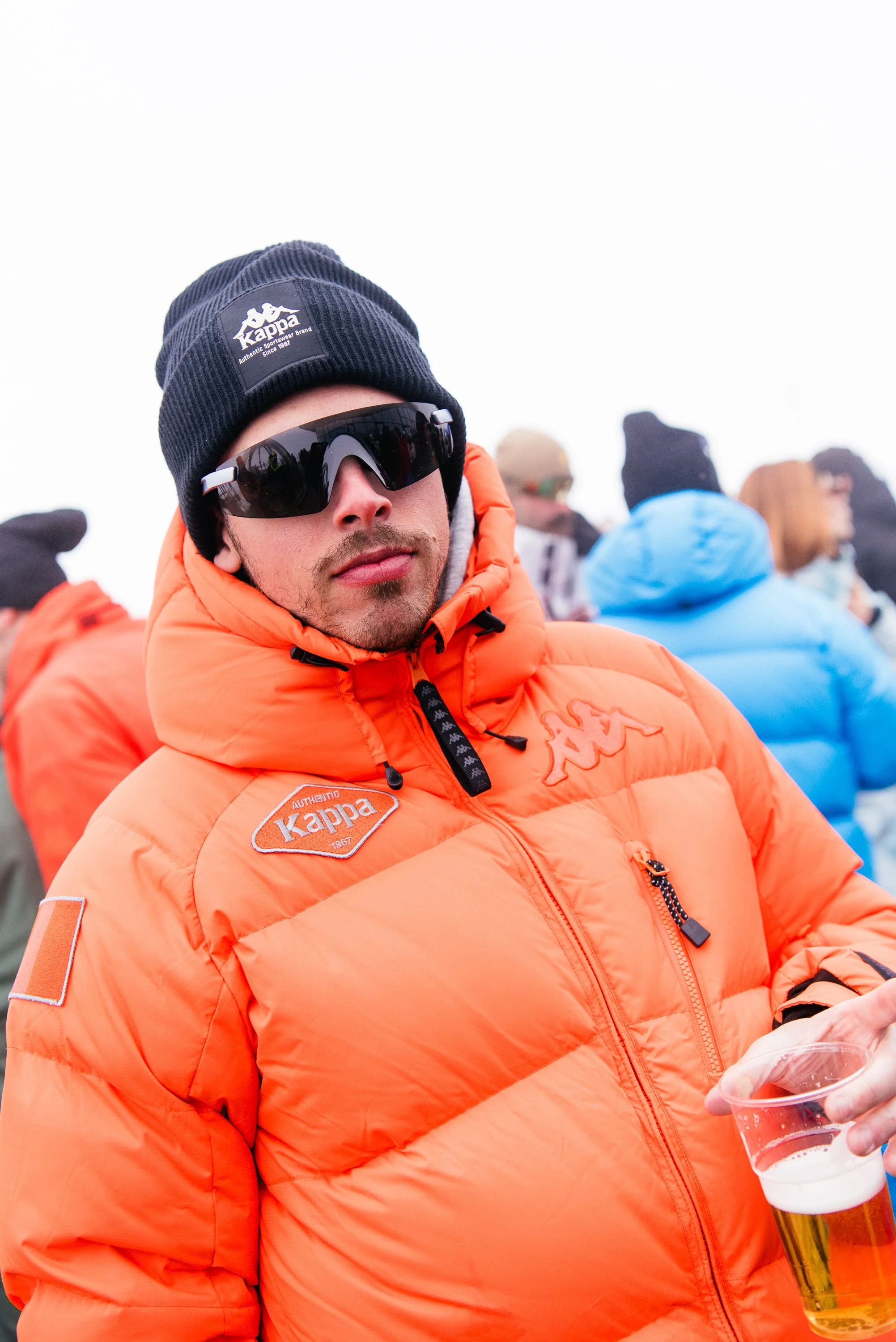 Un uomo con giacca arancione, cappello nero, occhiali da sole, e bicchiere di birra in mano.