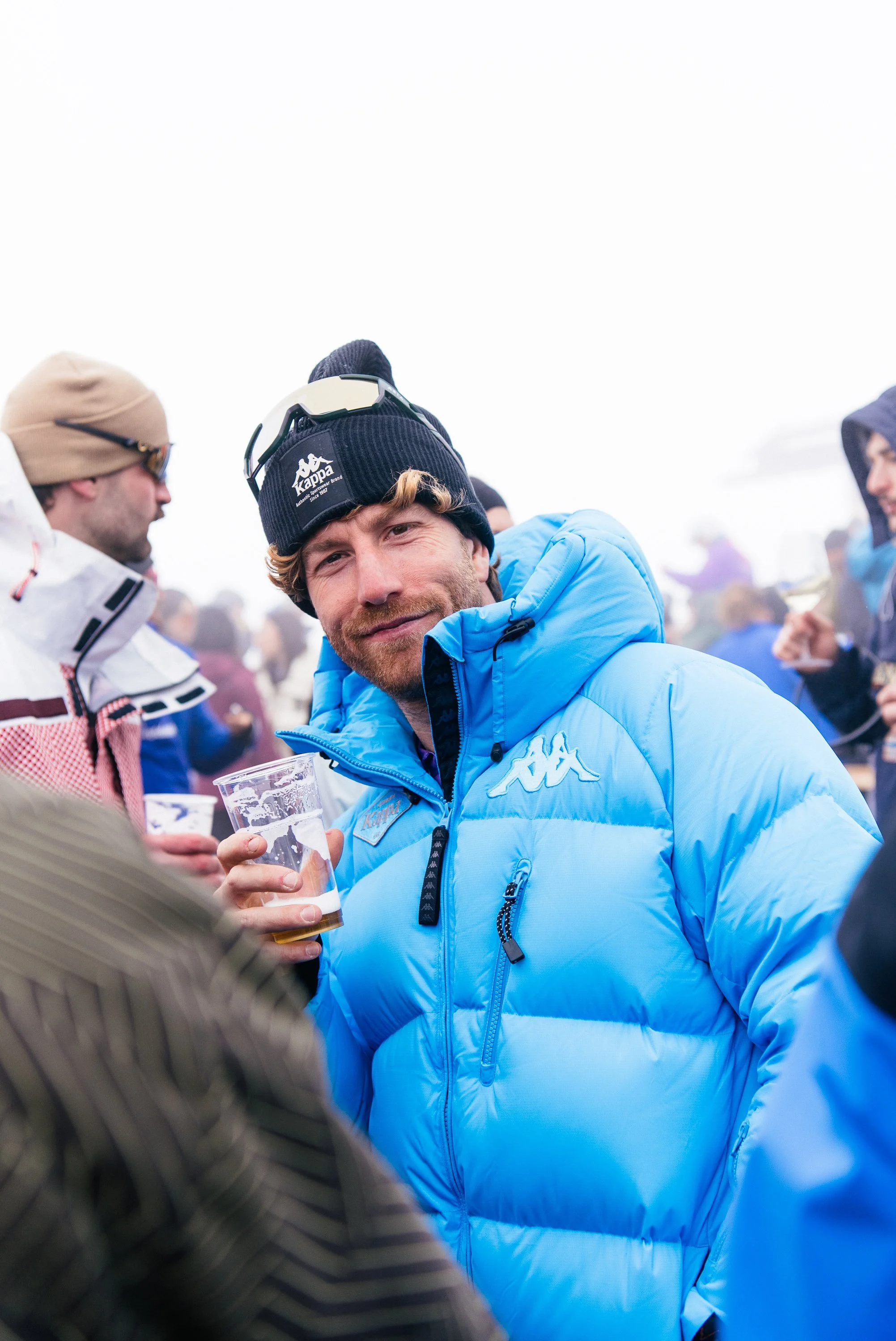 Un uomo con giacca blu e cappellino nero sorride mentre tiene un bicchiere di birra, circondato da altre persone vestite in abbigliamento da neve in un contesto all'aperto.