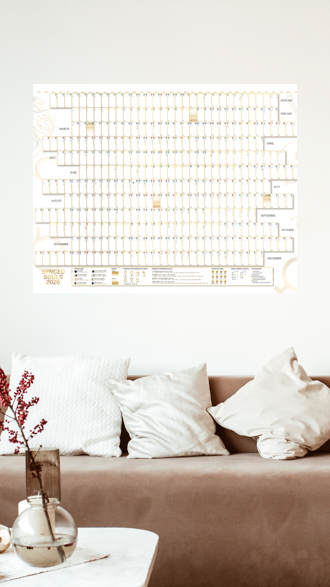 A wall calendar for the year 2026 titled 'Synced Souls' hangs above a beige sofa with three white pillows and a cream-colored pillow. In the foreground, a white tabletop features a clear glass vase with red berries.
