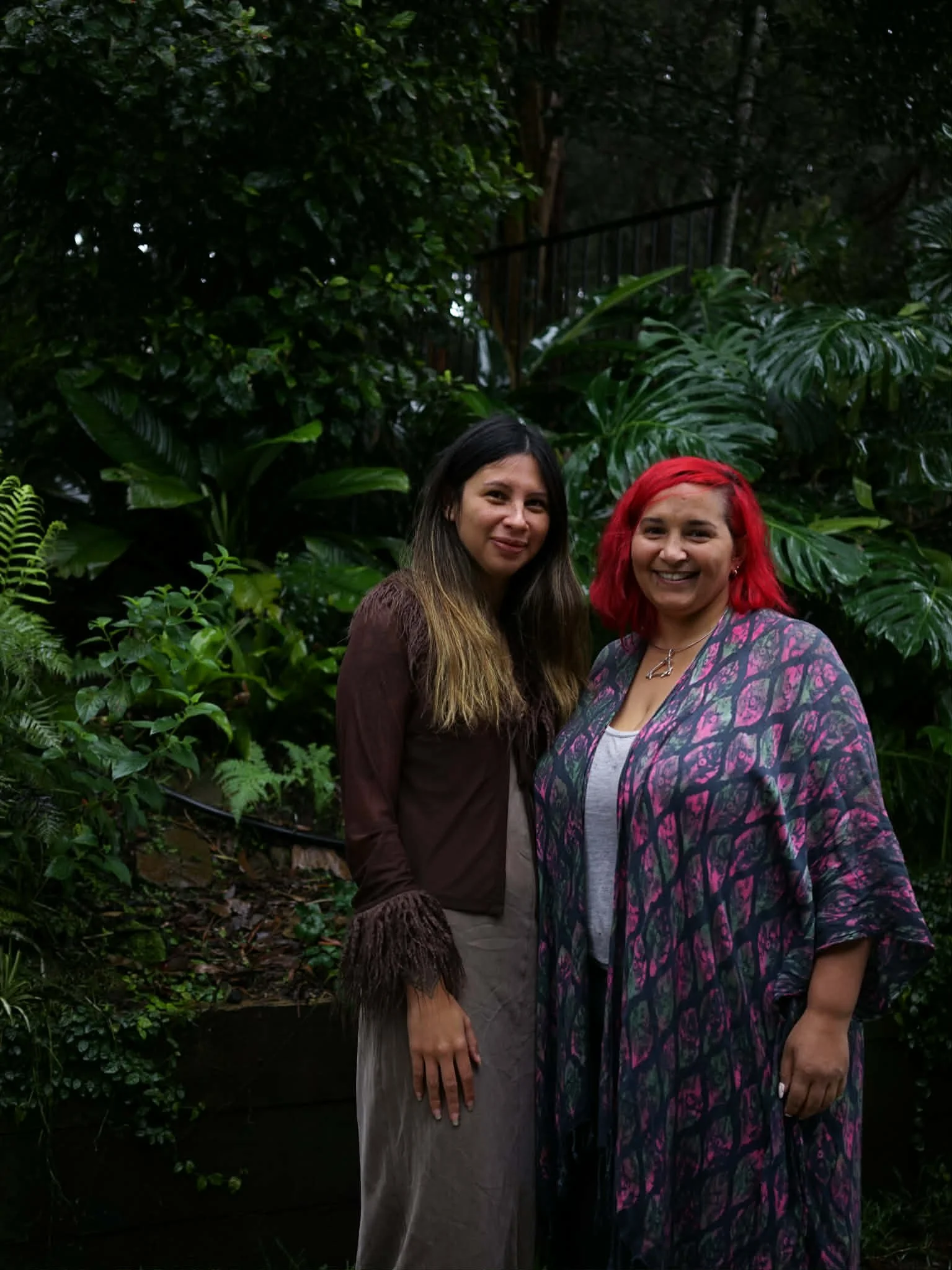 Two women standing outdoors in front of lush green plants, smiling at the camera.
