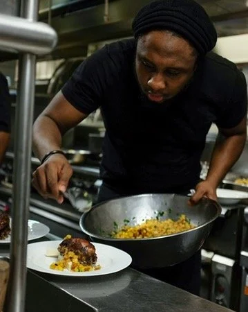 Chef garnishing a plated dish with a finishing touch in a professional kitchen.