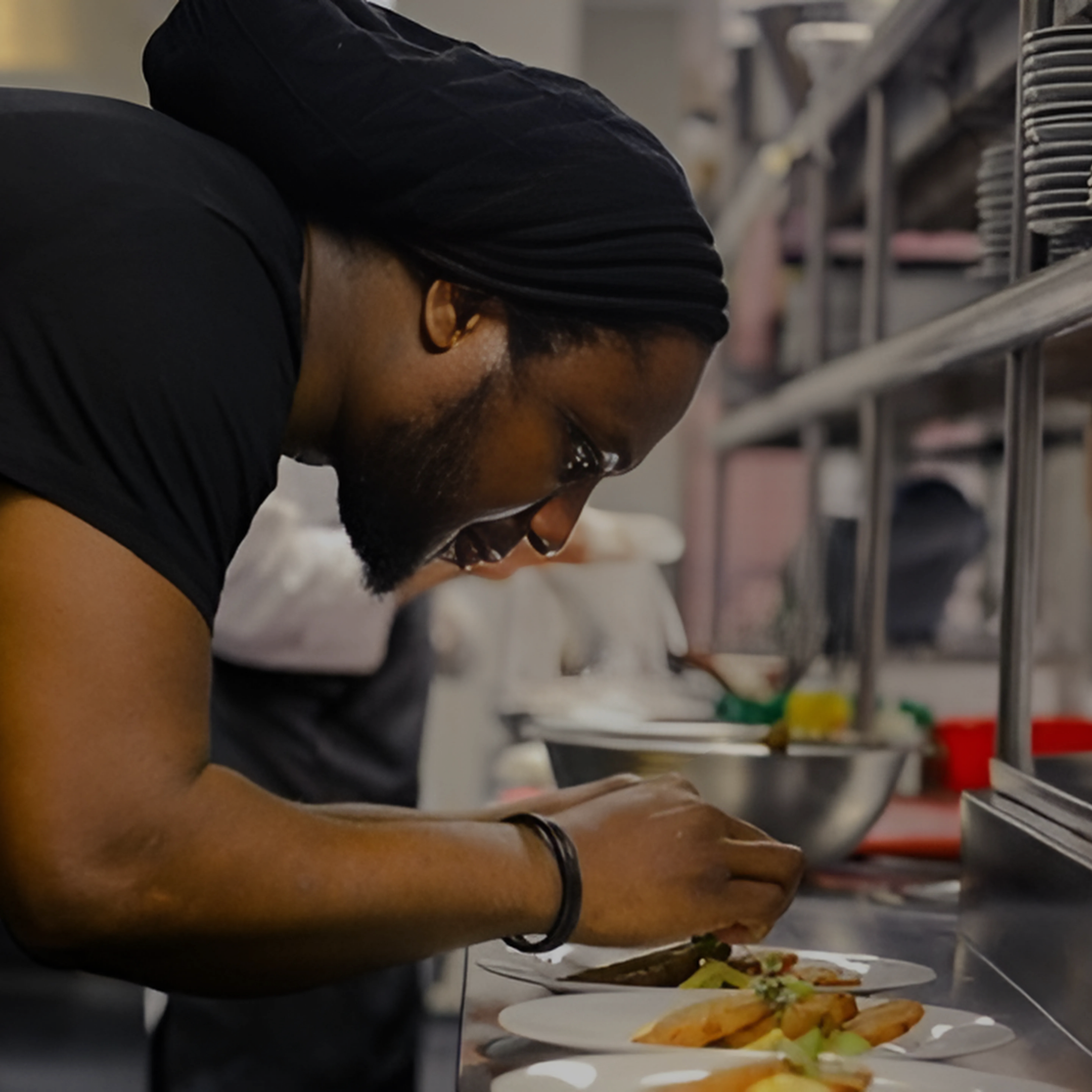 A chef plating food in a professional kitchen, wearing glasses and a black head covering, with bowls and kitchen utensils in the background.