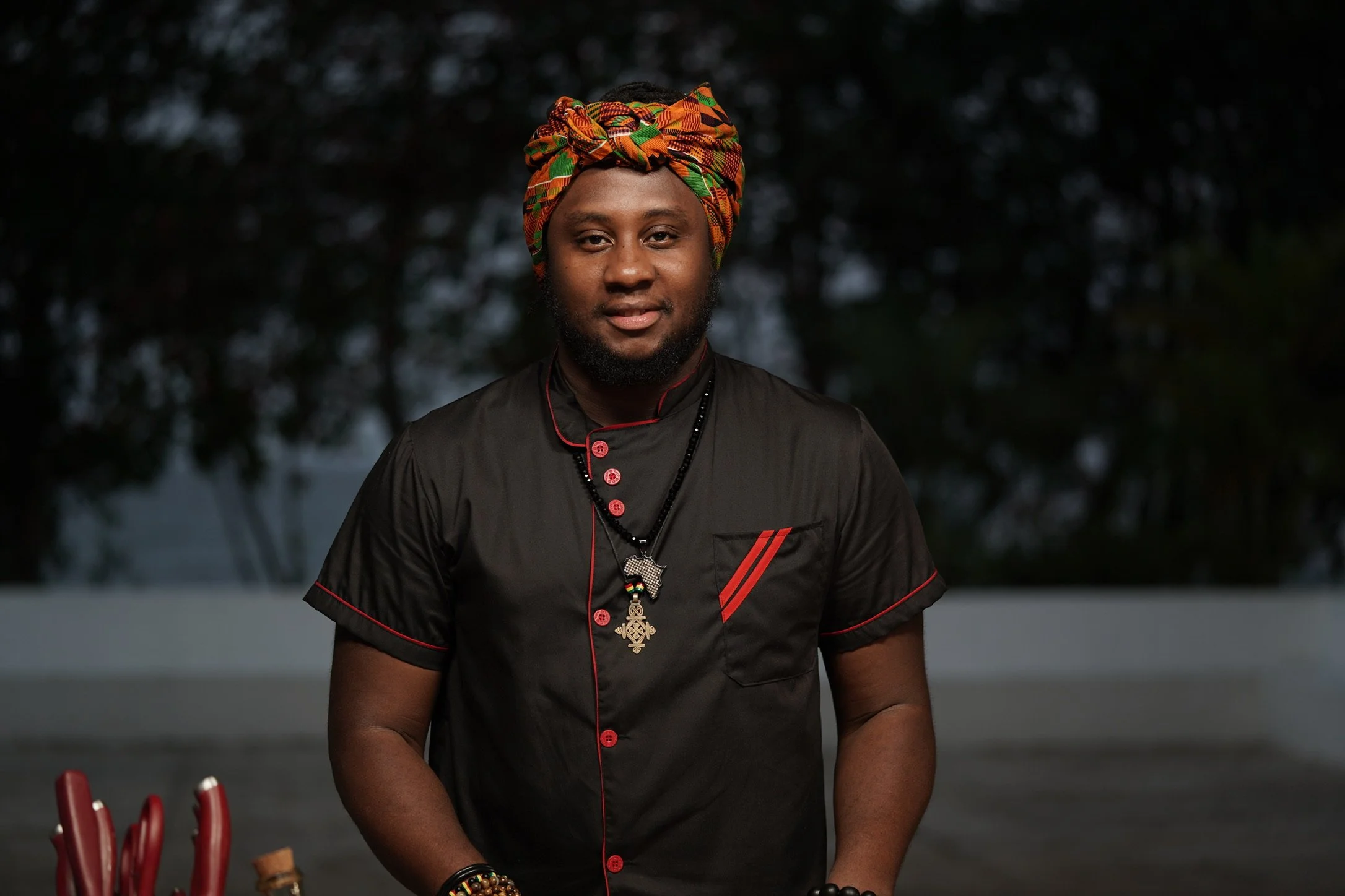 A man wearing a black shirt with red accents and colorful headwrap, standing outdoors during evening hours.
