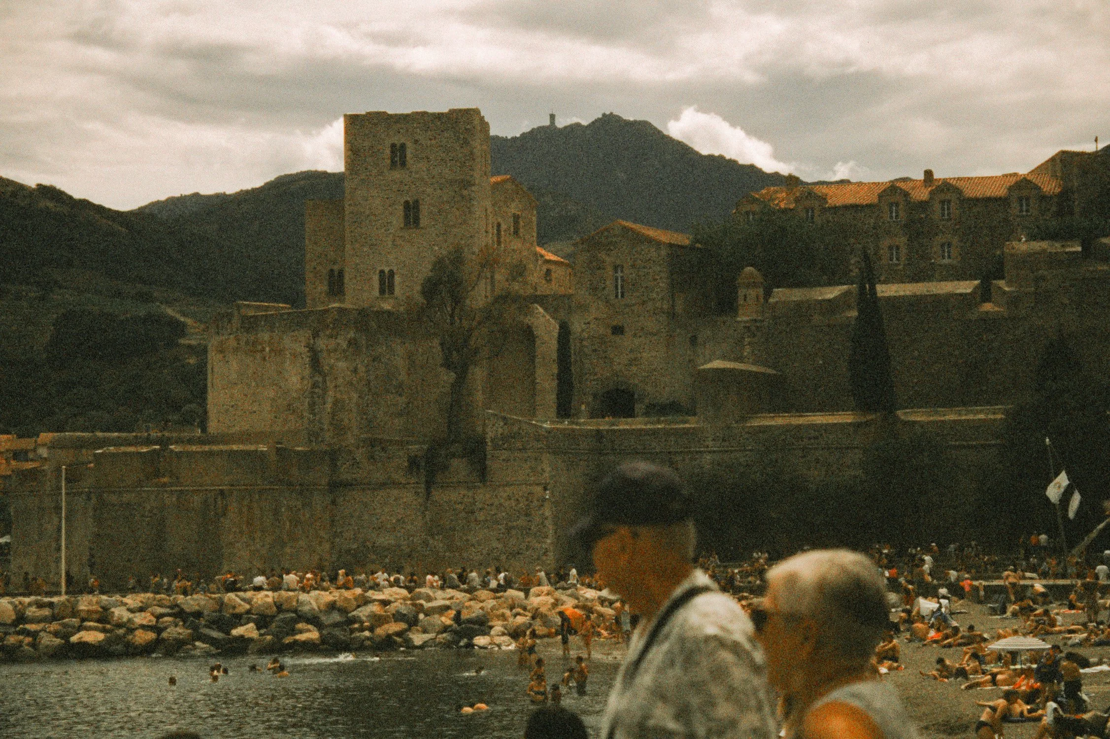 A castle with stone walls and towers on a hill overlooking a crowded beach with many people swimming and relaxing, mountains in the background, and an overcast sky.