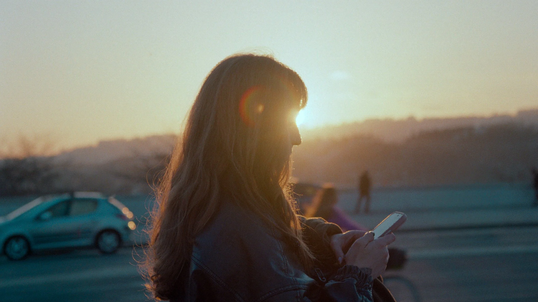 A woman with long hair looking at her phone during sunset at a parking lot with cars in the background.