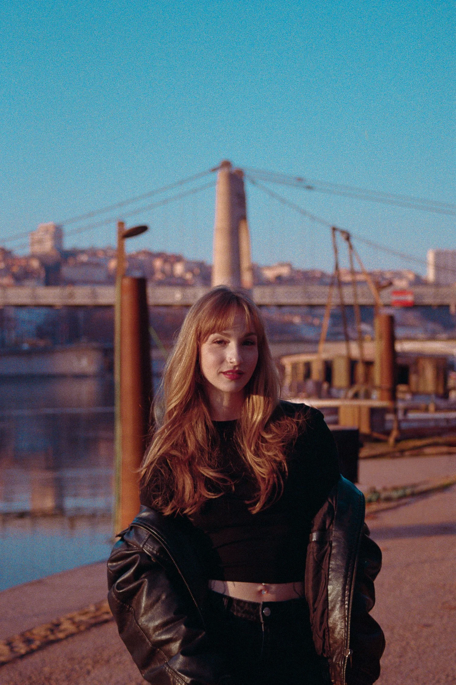 A young woman with long, wavy red hair standing outdoors near water, wearing a black crop top, black jeans, and a black leather jacket draped over her shoulders, with a bridge and cityscape in the background during sunset.
