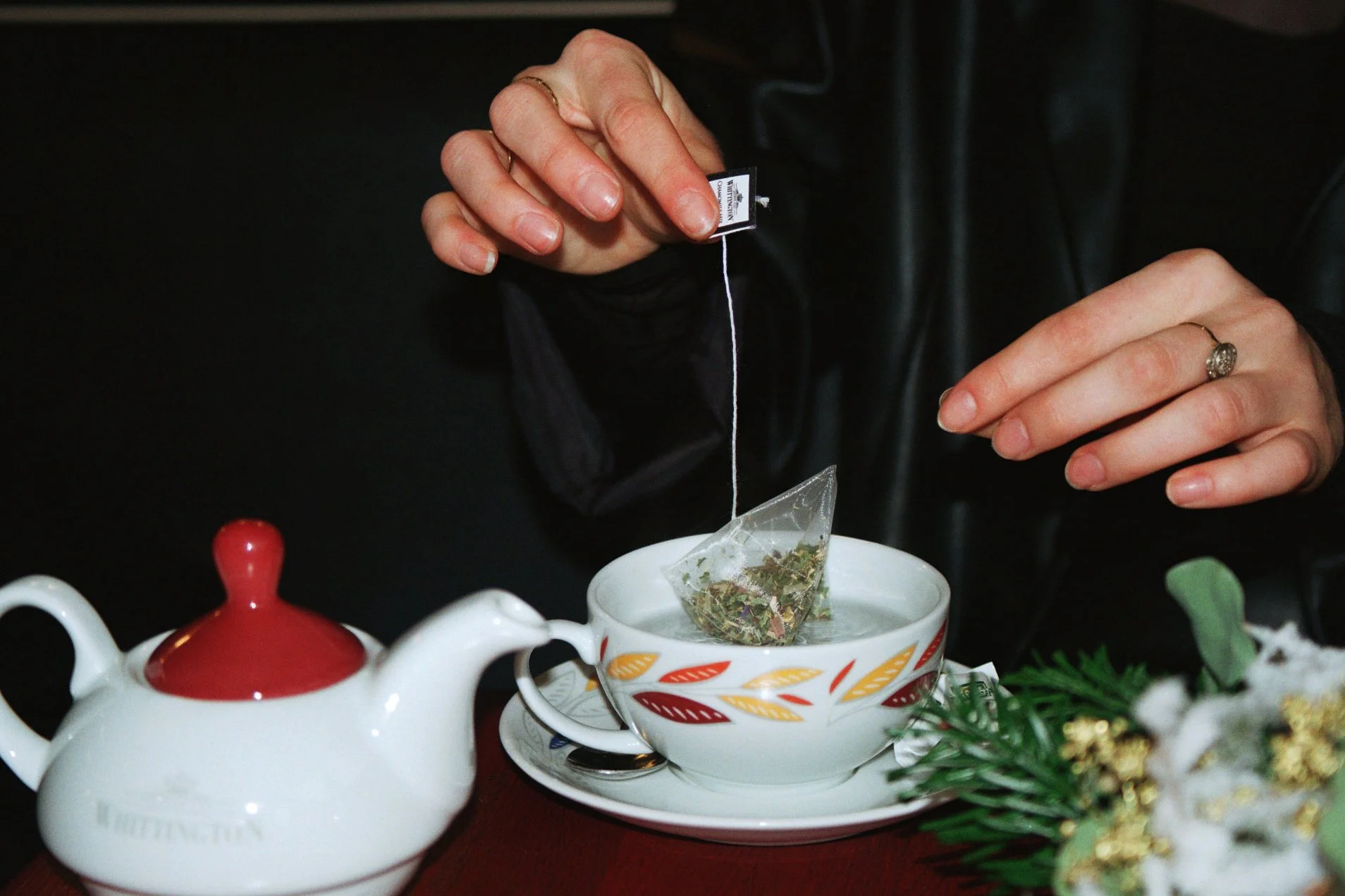 Person holding a tea bag about to steep it in a white patterned teacup on a saucer, with a white teapot nearby, and holiday decorations on the table.