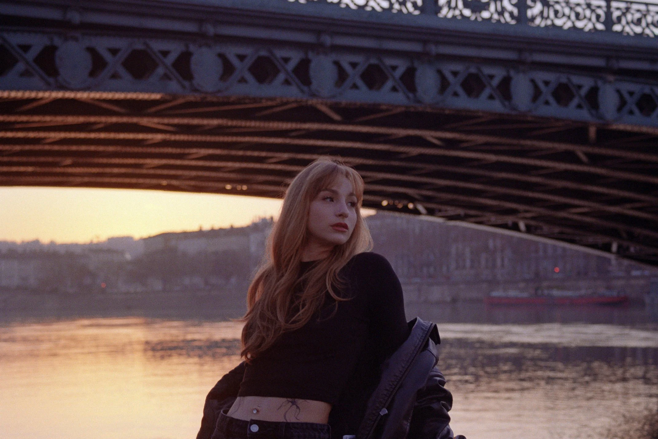 A young woman with long red hair standing under a bridge by the river at sunset, wearing a black top and jeans, with a jacket hanging off her shoulder.