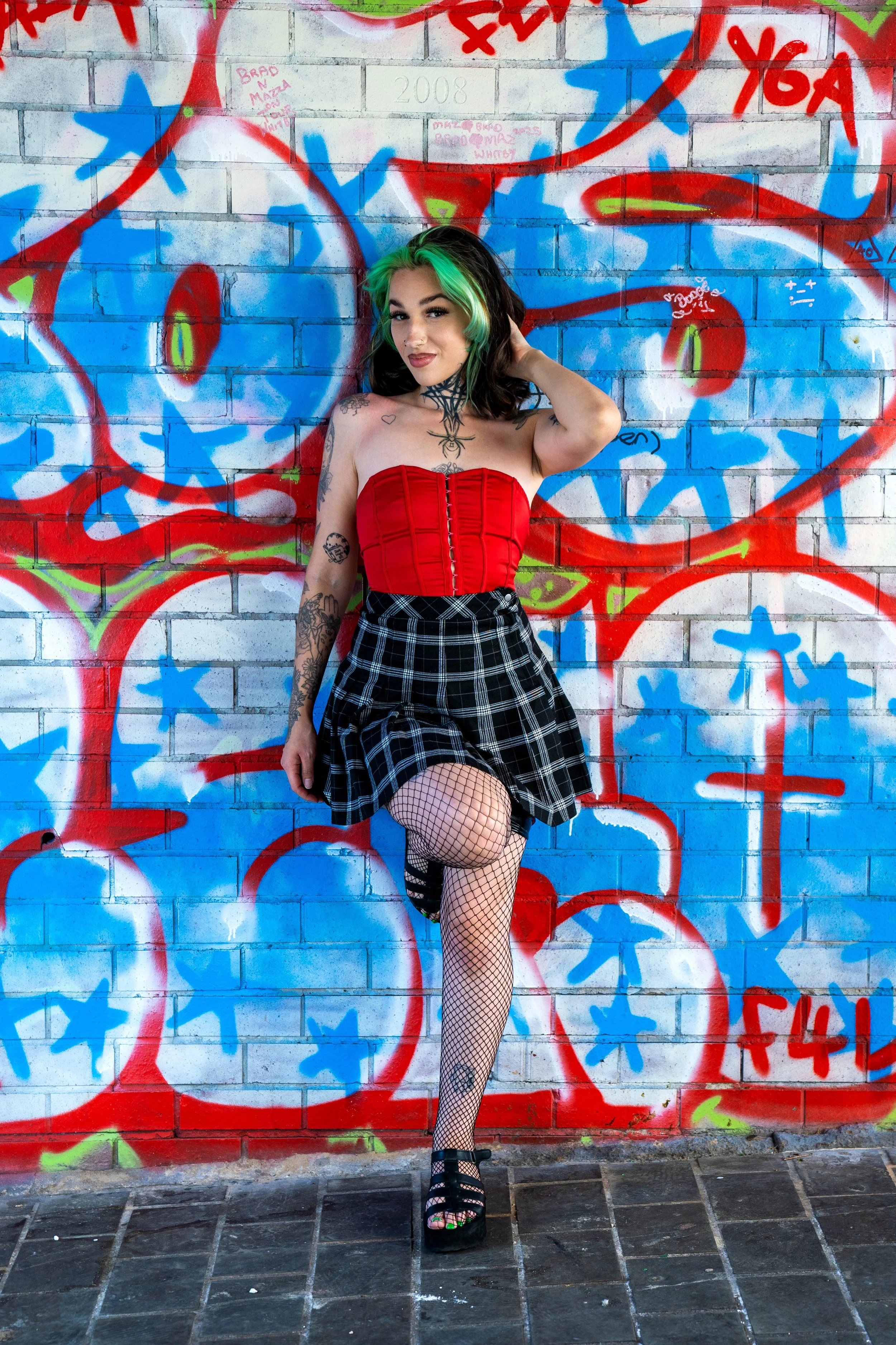 Young woman with dark hair and green highlights, wearing a red strapless top, plaid skirt, fishnet stockings, and black platform heels, standing against a graffiti-covered brick wall.