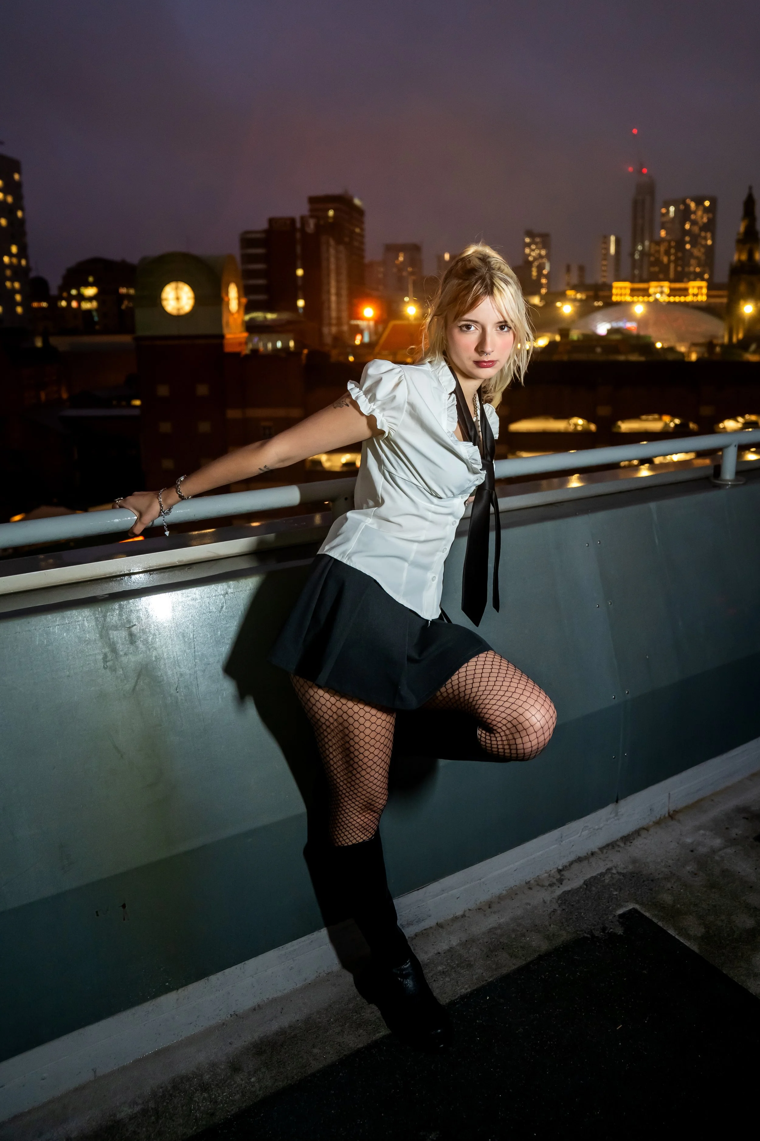 A woman in a white blouse, black skirt, fishnet stockings, and black boots posing on a rooftop at night with city lights and skyscrapers in the background.