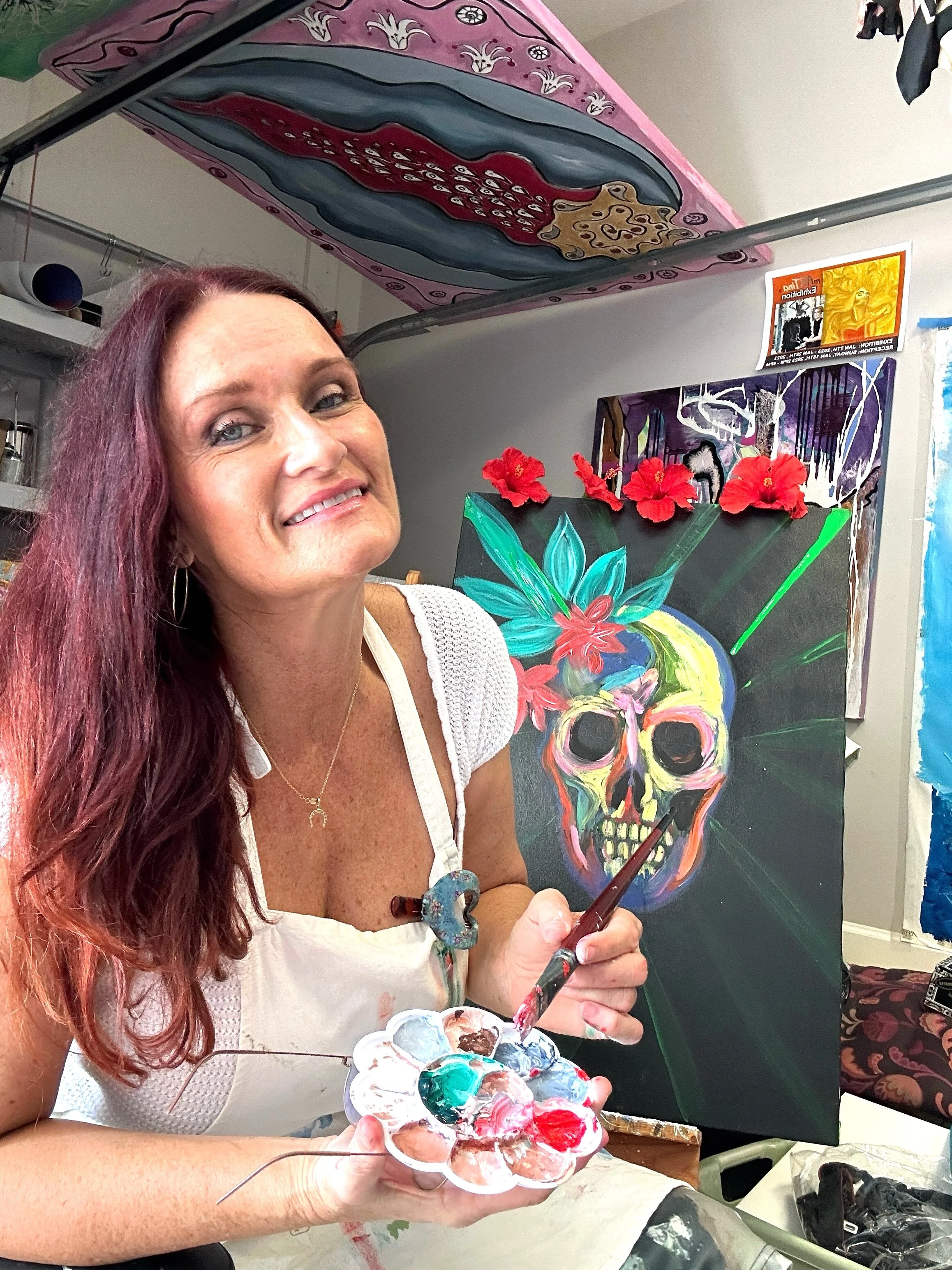 A woman with long red hair smiling and holding a palette with various colors of paint, working on a painting of a colorful skull adorned with flowers on a black background in an art studio.