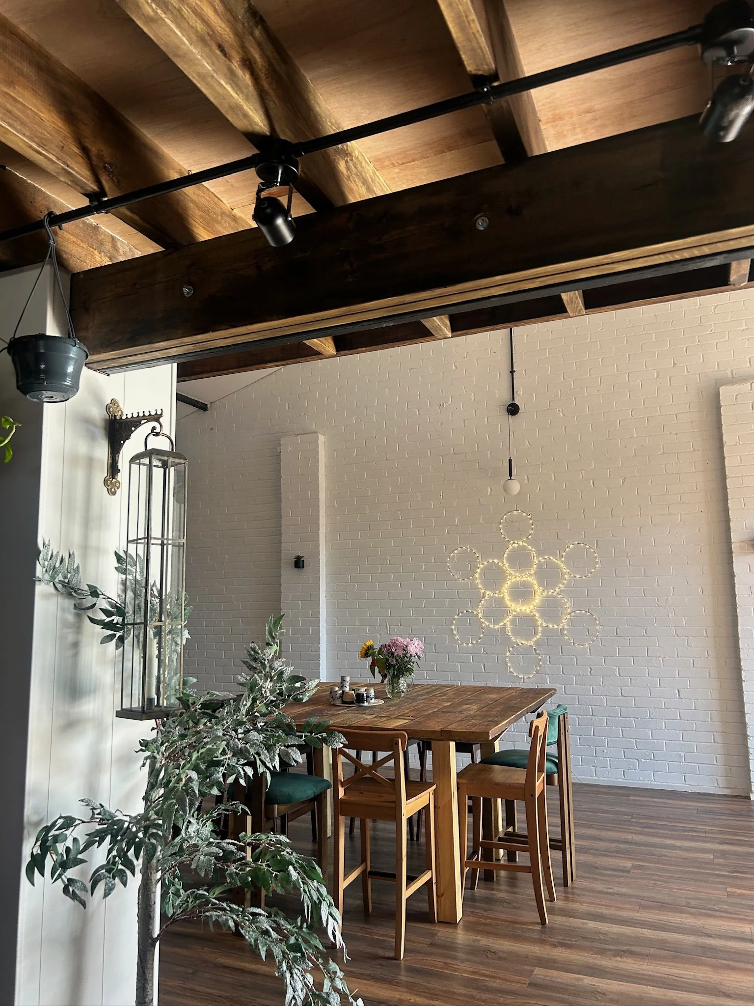 A modern dining area with a wooden table surrounded by four chairs, a bouquet of flowers, and decorative lighting on the white brick wall.