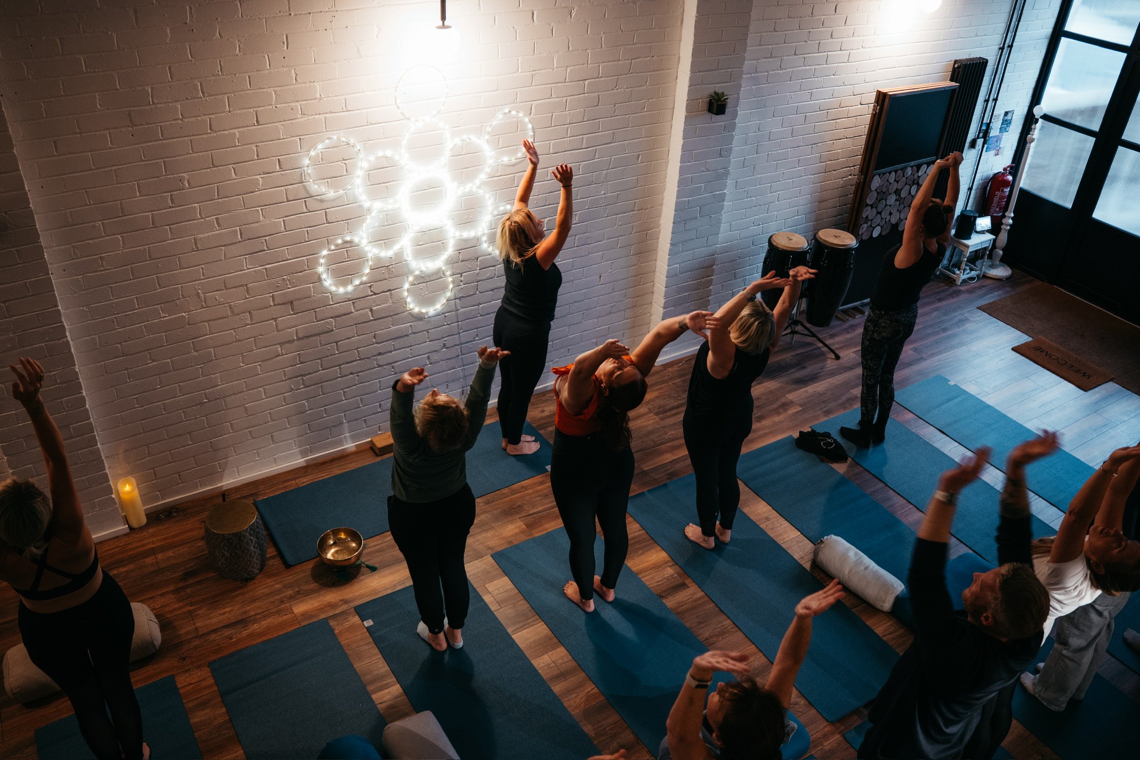 Group of people practicing yoga in a studio with white brick walls, blue mats, and a neon light decoration on the wall.