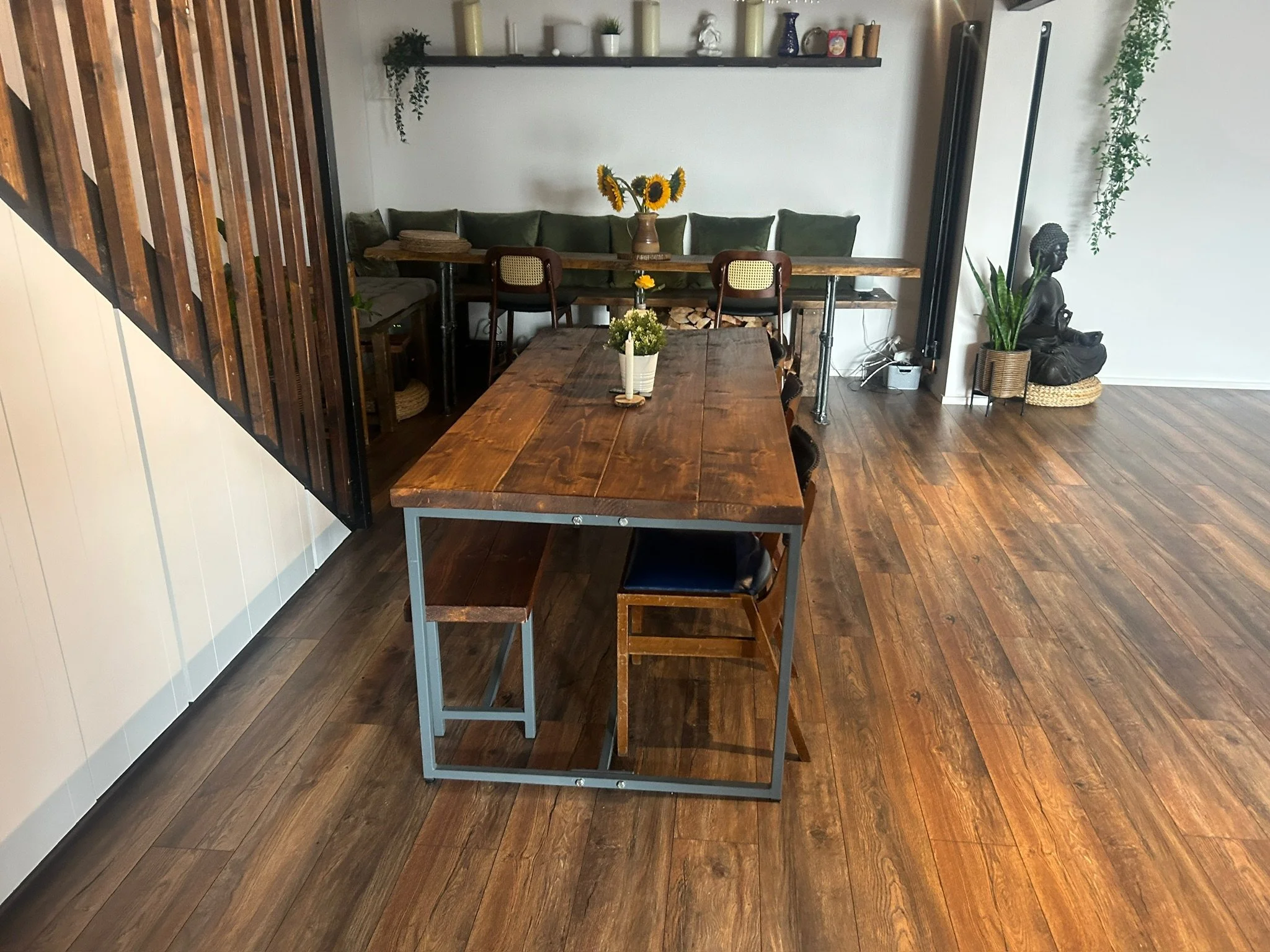 A cozy dining area with a wooden table set for several people, on a dark wood floor. In the background, there is a long bench with green cushions and a vase with sunflowers on a wall-mounted shelf. To the right, a decorative statue of a sitting Buddh