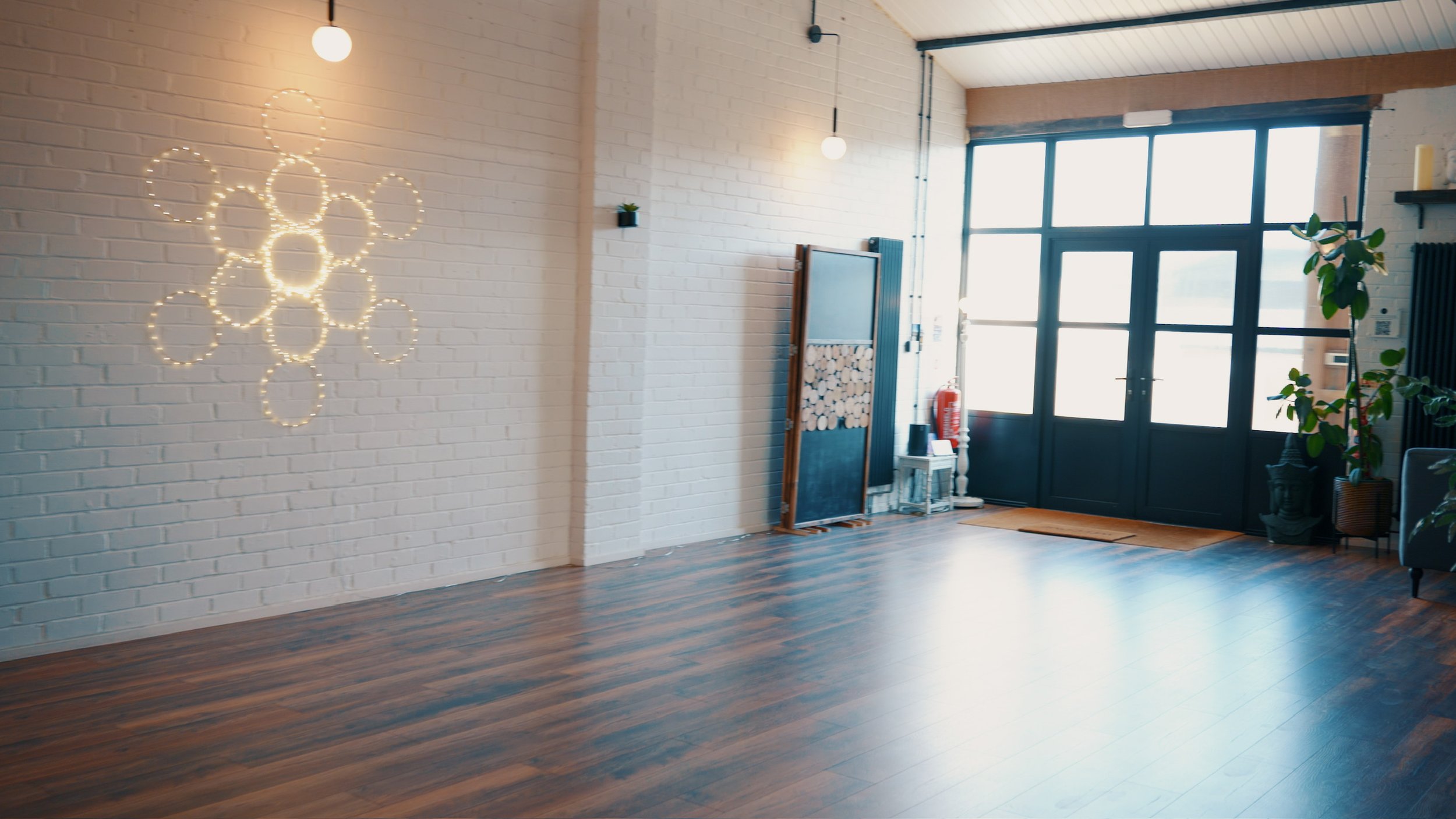 Empty room with a white brick wall, wooden floor, large window, some plants, and decorative lights on the wall.