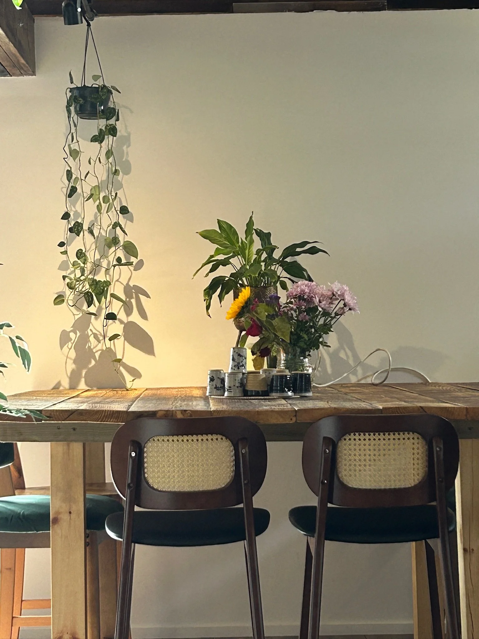 A wooden dining table with two chairs, decorated with bouquets of flowers in vases, and a hanging potted plant on the wall behind.