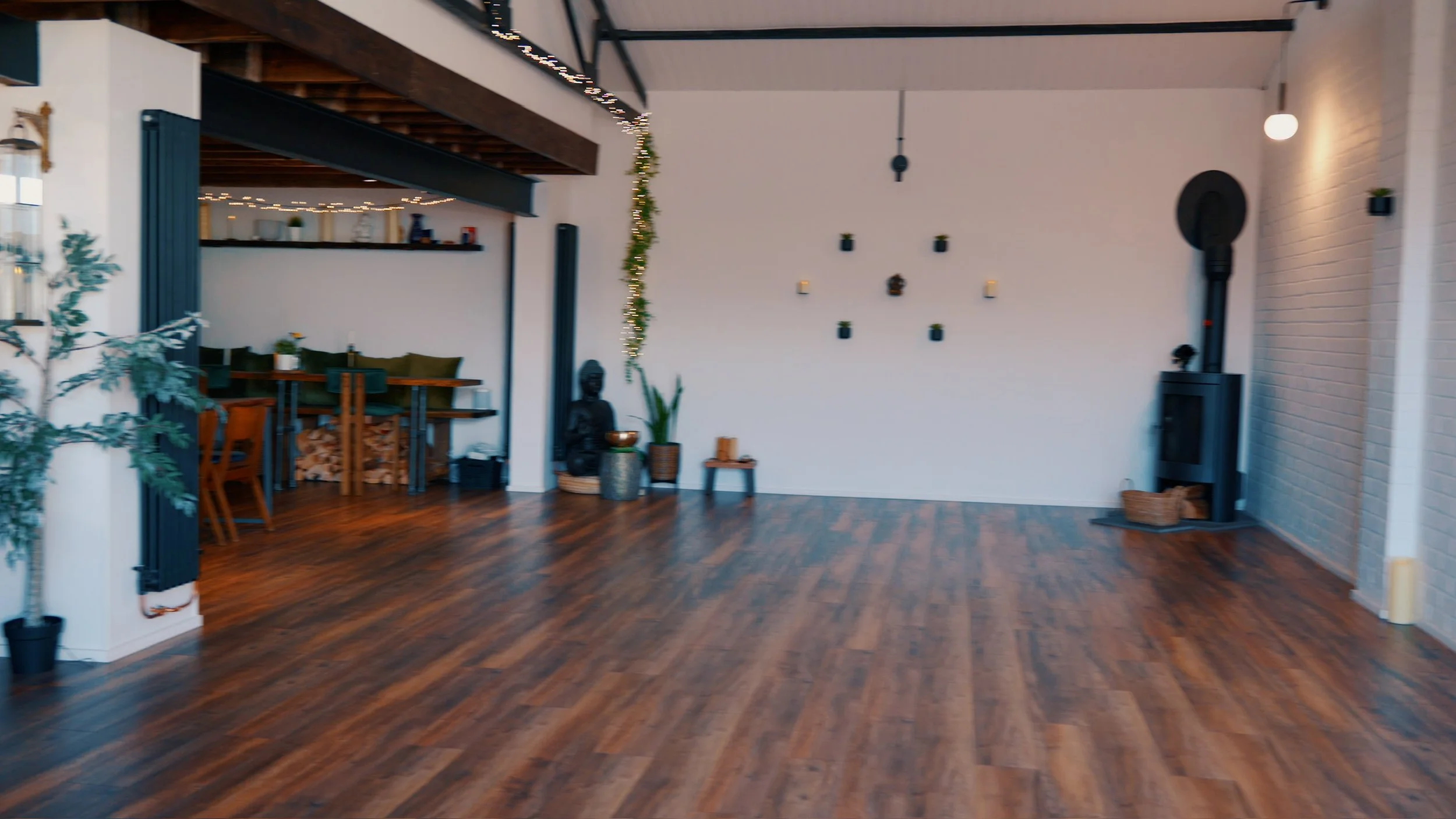 An empty living room with wood flooring, a white brick wall, and minimalist decor including wall-mounted candles, potted plants, and a black stove with a chimney.