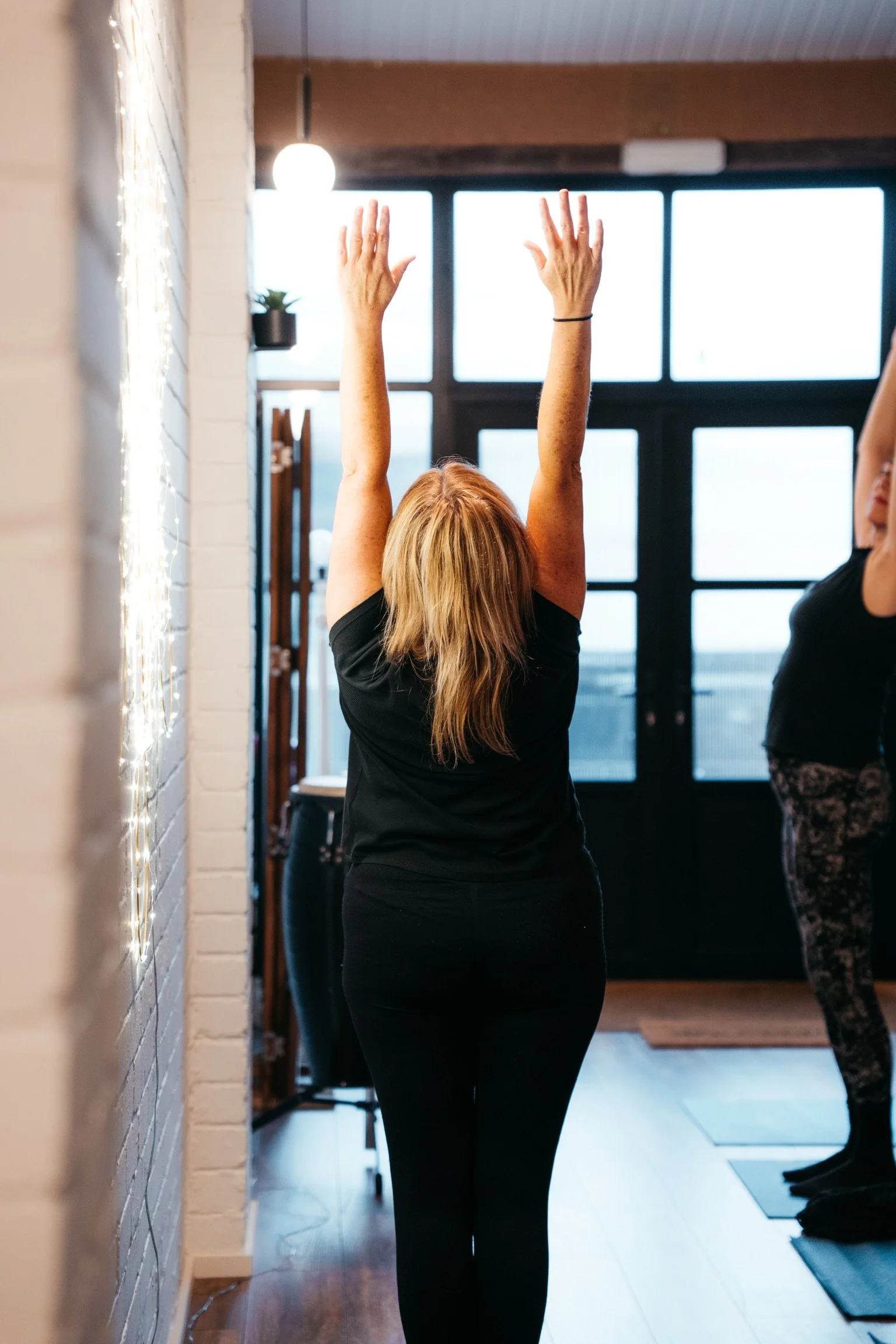 Woman in black exercise attire doing a stretch with arms raised above head in a yoga or workout studio, with another person partially visible in the background.