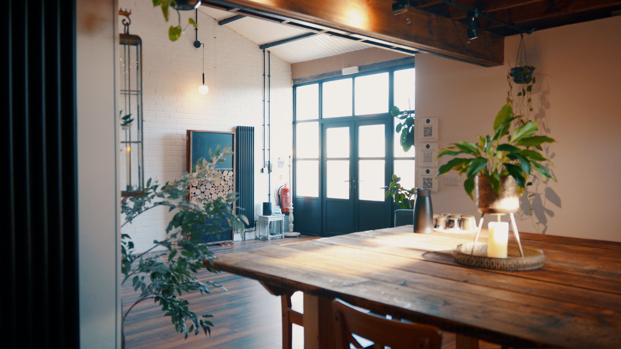 Interior of a cozy room with a wooden table in the foreground adorned with a potted plant, candles, and a black pitcher. In the background, large windows let in natural light, and there are plants hanging on the wall, a framed art piece, a fire extin