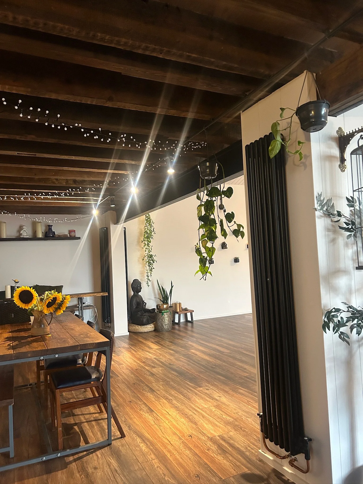 Interior of a modern living room with wooden floors and ceiling, decorative plants, a sculpture of Buddha, and string lights hanging from the ceiling.