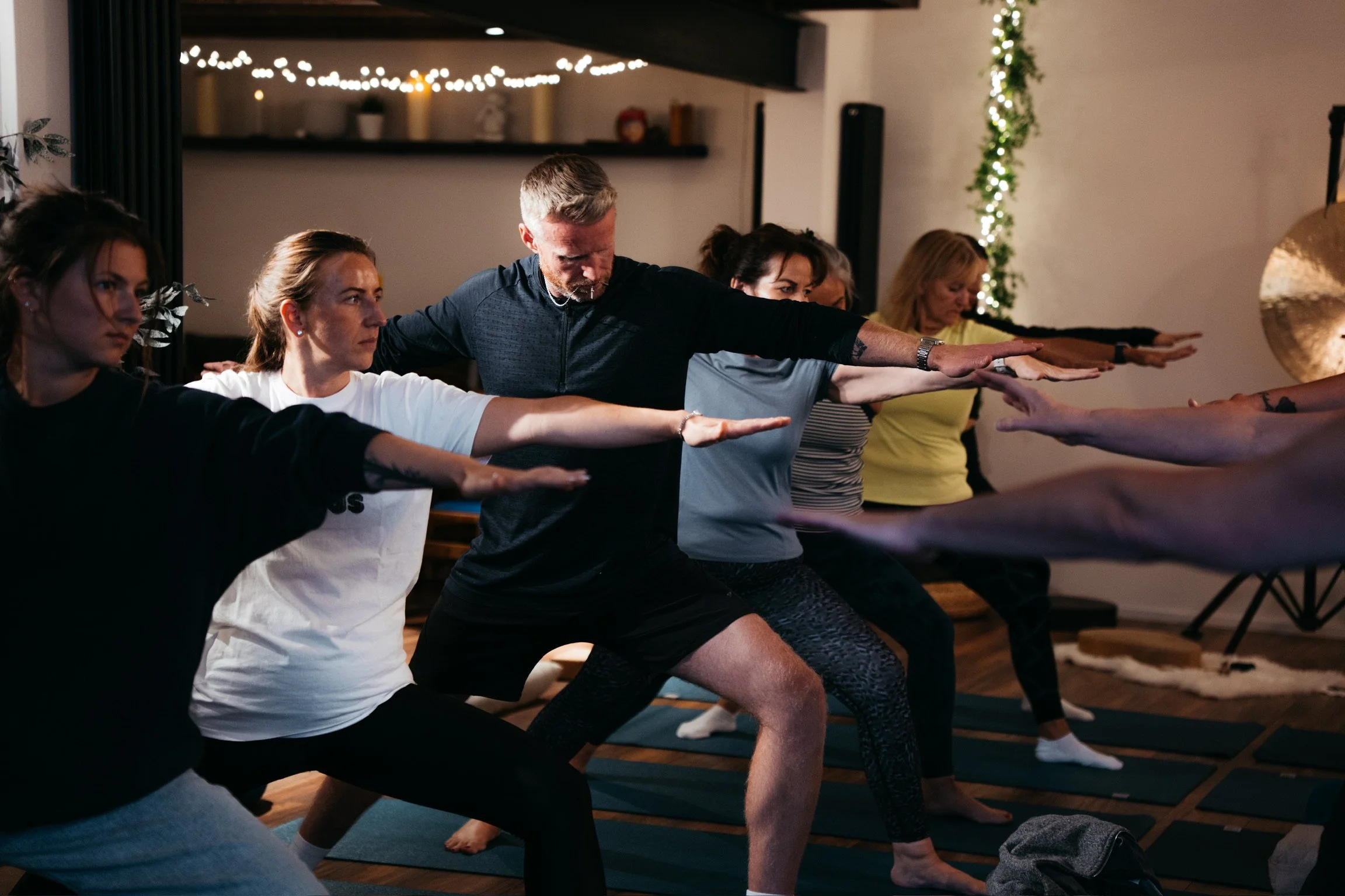 Group of people practicing yoga in a studio, extending arms forward, with festive string lights and a decorated Christmas tree in the background.