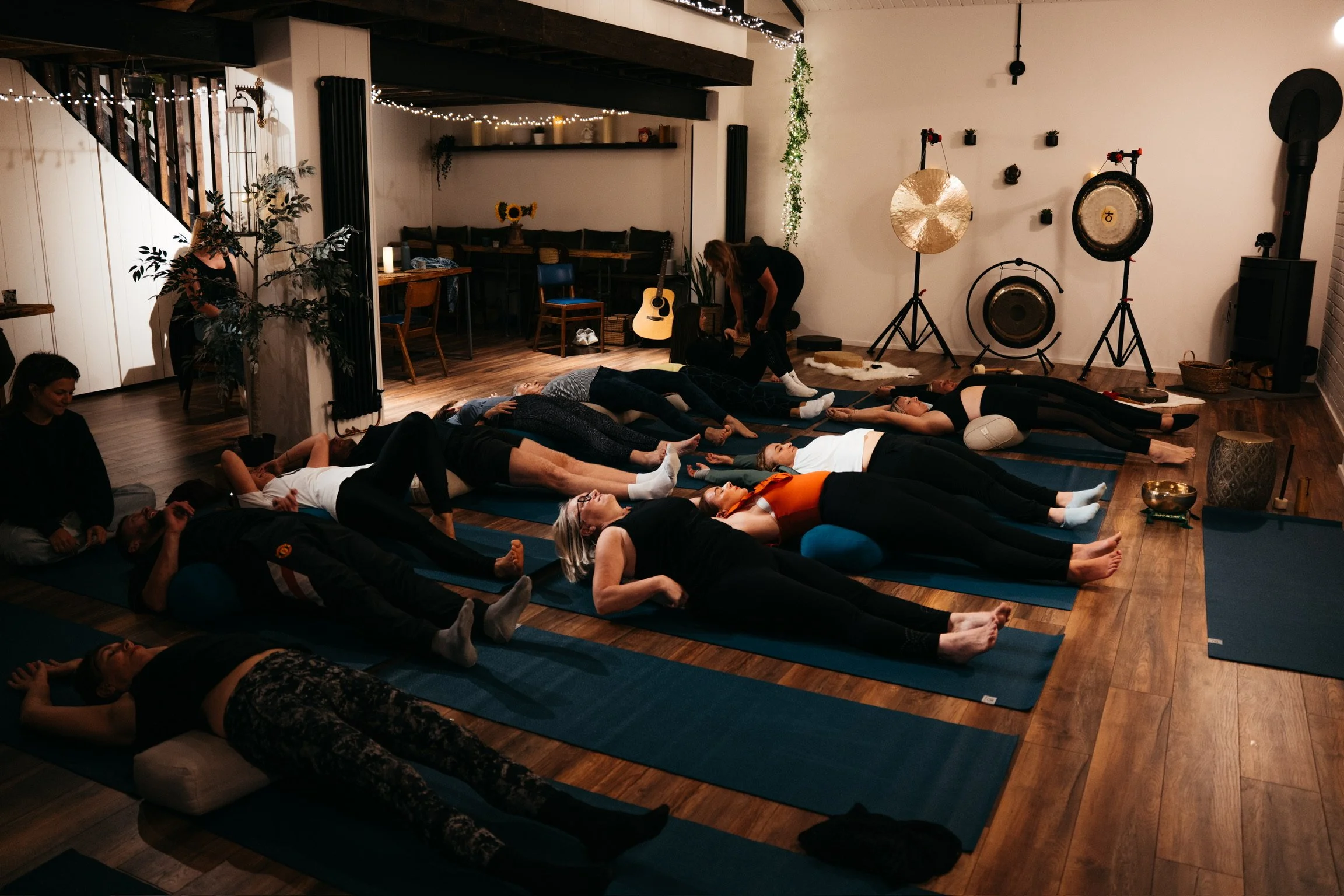 People participating in a yoga or meditation class lying on mats in a cozy, dimly lit room with musical instruments and decorative gongs in the background.