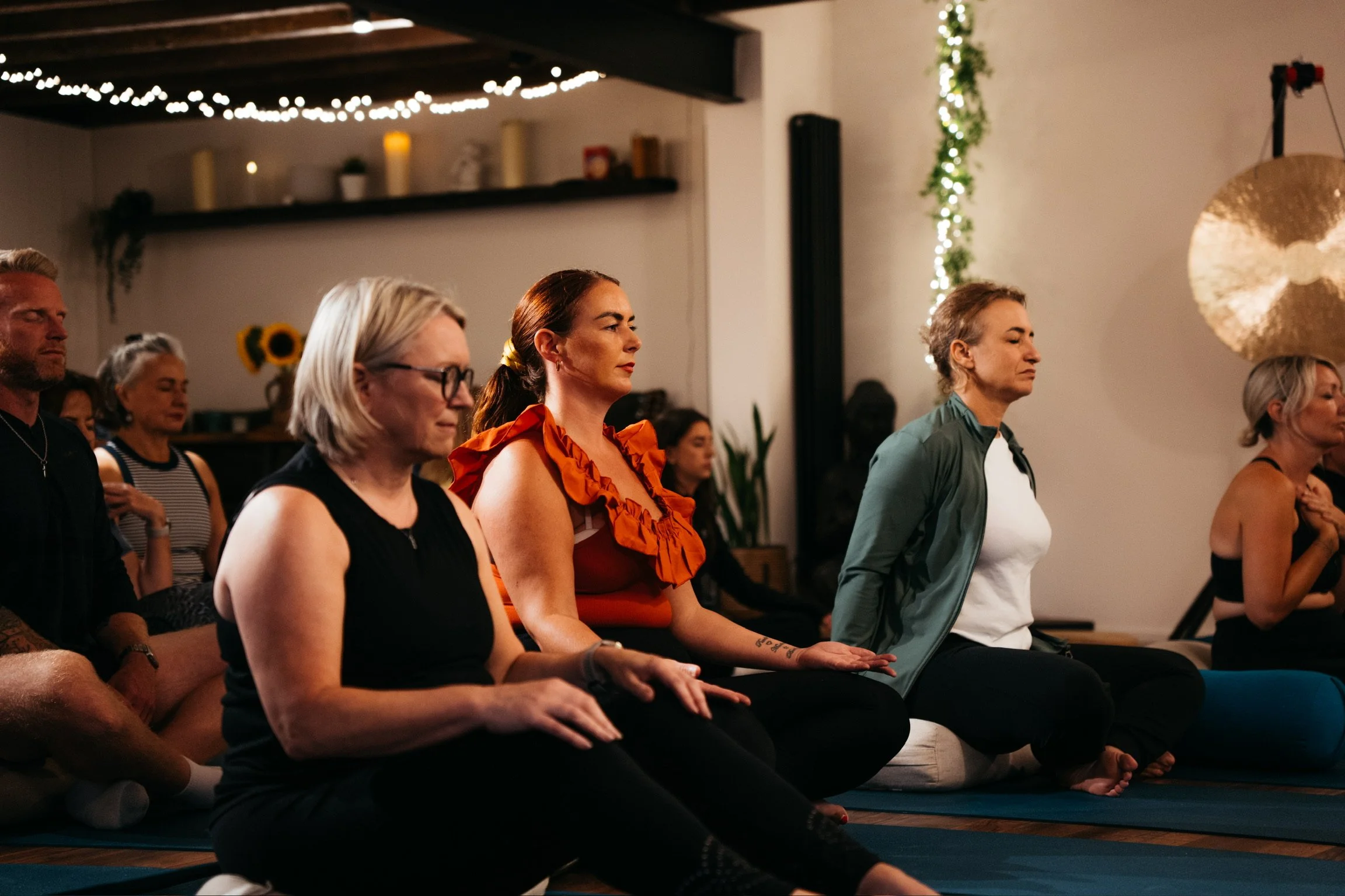 People sitting on yoga mats in a meditation or yoga class, with eyes closed and hands resting on knees, in a cozy indoor space decorated with string lights, candles, sunflowers, and a large gong.